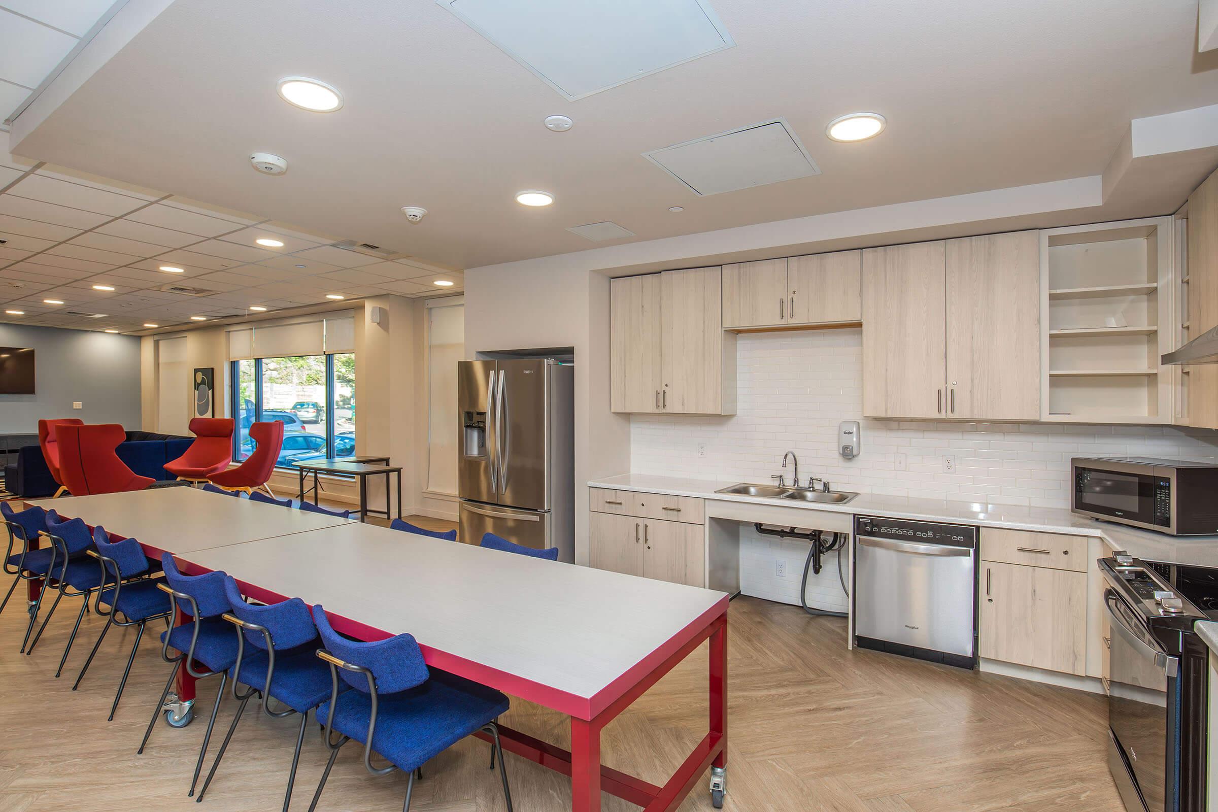 A spacious kitchen and dining area featuring a long table with blue chairs, a modern kitchen with stainless steel appliances, and a cozy seating area with red lounge chairs. Large windows allow natural light to fill the space, creating a welcoming atmosphere for gatherings.