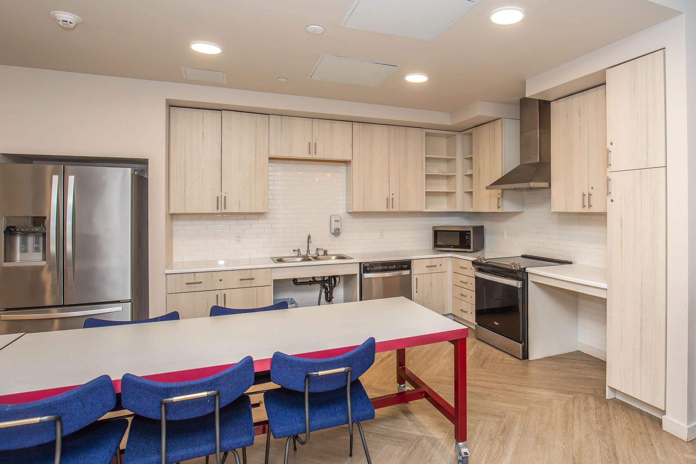 Modern kitchen with light wood cabinets, stainless steel appliances including a refrigerator and oven, and white subway tile backsplash. A central island with a gray tabletop and red legs is surrounded by blue chairs. Well-lit space with recessed lighting and a contemporary design.