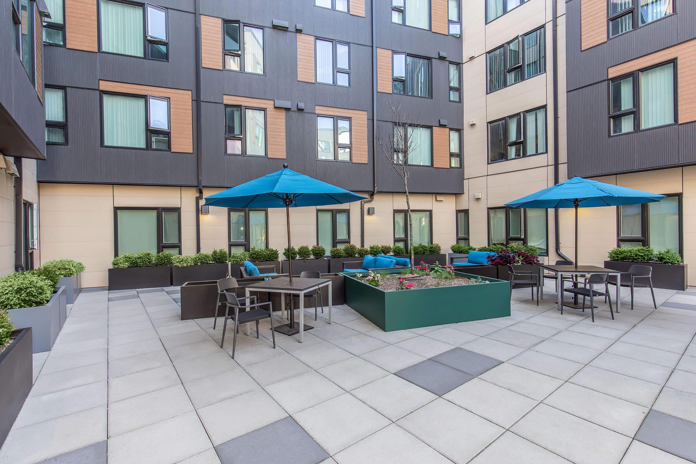 A modern outdoor courtyard featuring seating areas with umbrellas, potted plants, and a central garden bed. The courtyard is surrounded by contemporary apartment buildings with large windows, creating a welcoming atmosphere for relaxation or social gatherings.