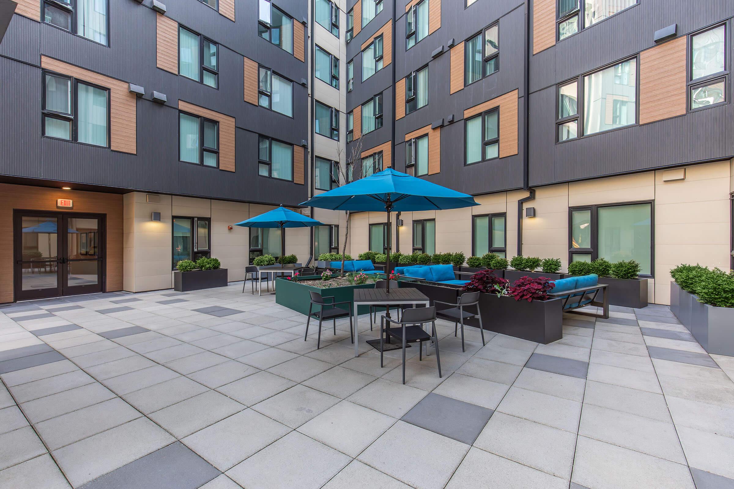 A modern outdoor courtyard featuring blue umbrellas, comfortable seating with cushions, and planters with greenery. The setting includes paved flooring and is surrounded by contemporary building architecture, creating an inviting communal space.