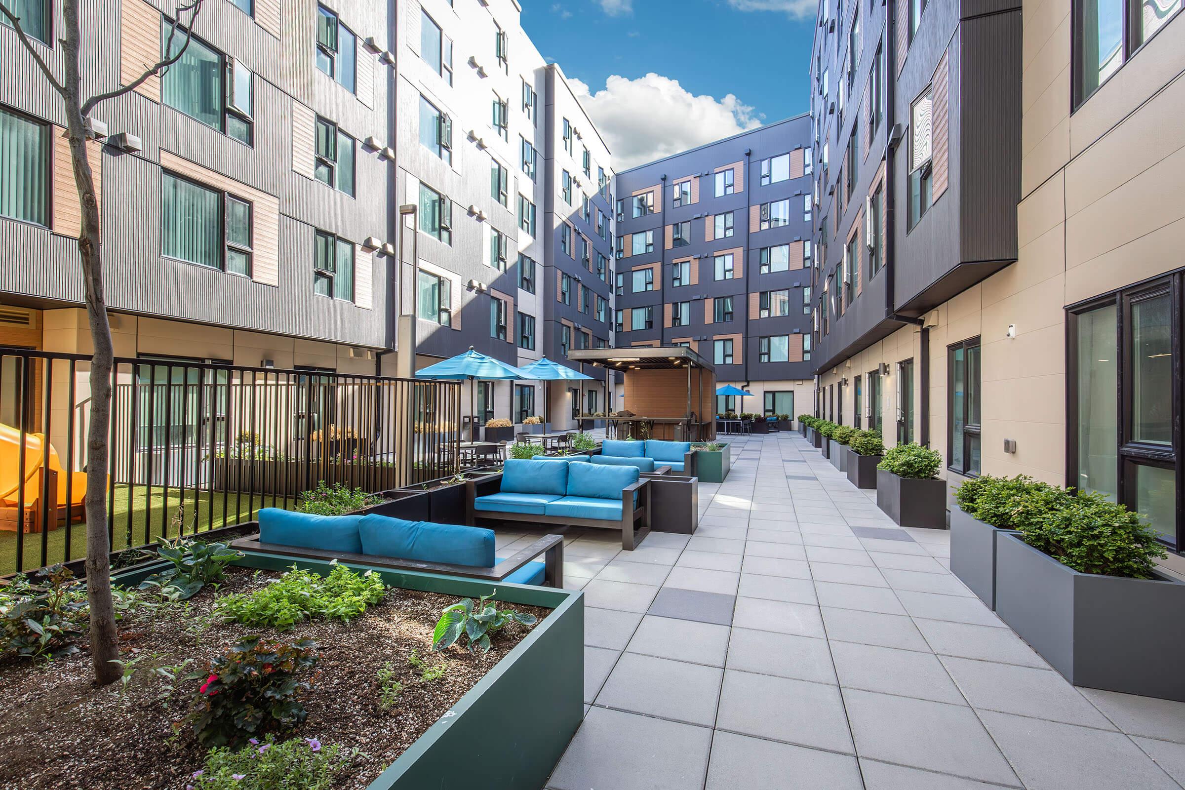 A modern outdoor courtyard featuring landscaped planters, blue seating areas, and patio umbrellas. Surrounding buildings have a contemporary design with windows reflecting the sky. The space is inviting, ideal for relaxation and social gatherings.