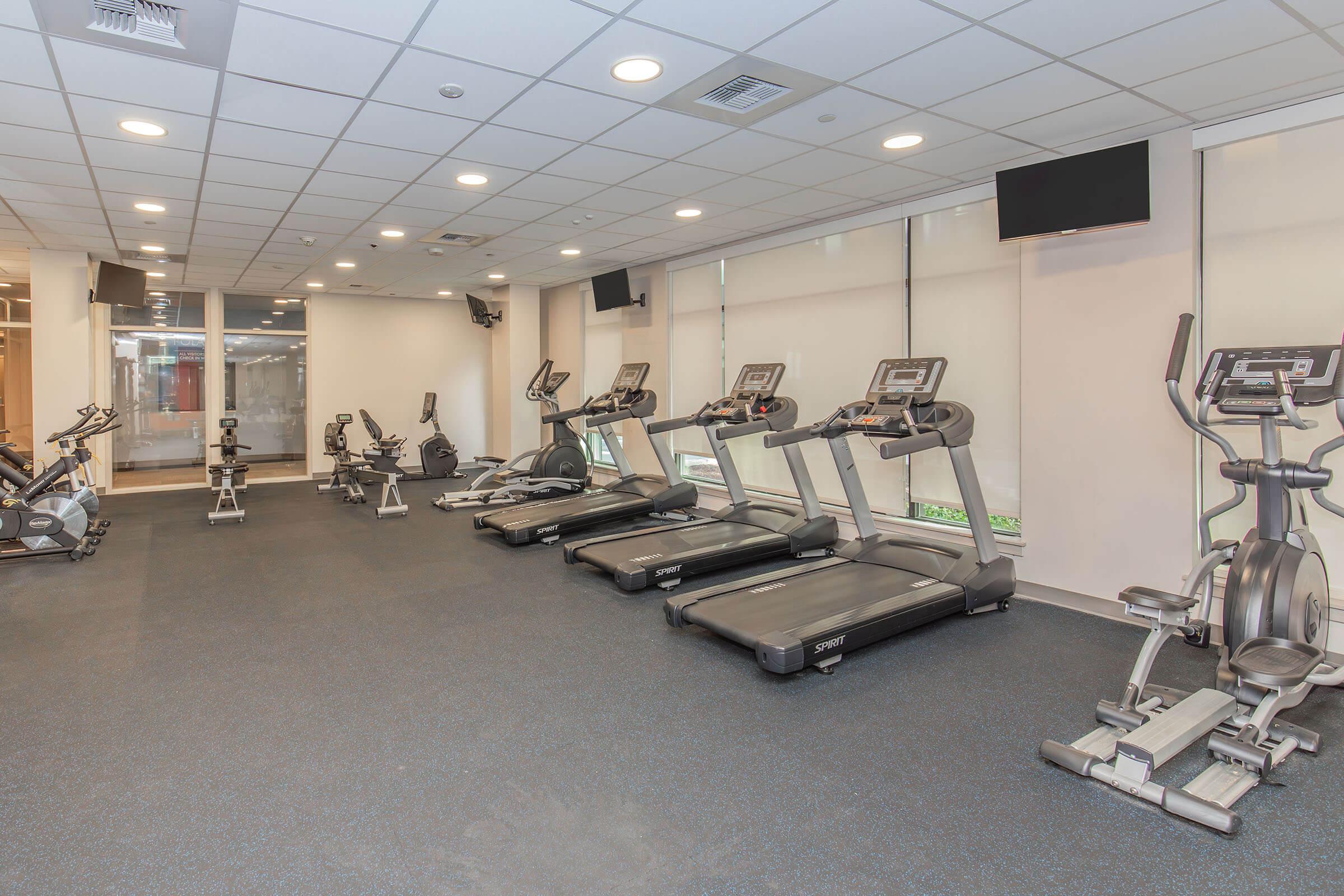 A spacious, modern gym interior featuring several treadmills and elliptical machines arranged neatly on a dark carpeted floor. Large windows allow natural light to enter the room, which is equipped with ceiling lights and wall-mounted TVs. The environment appears clean and well-maintained.