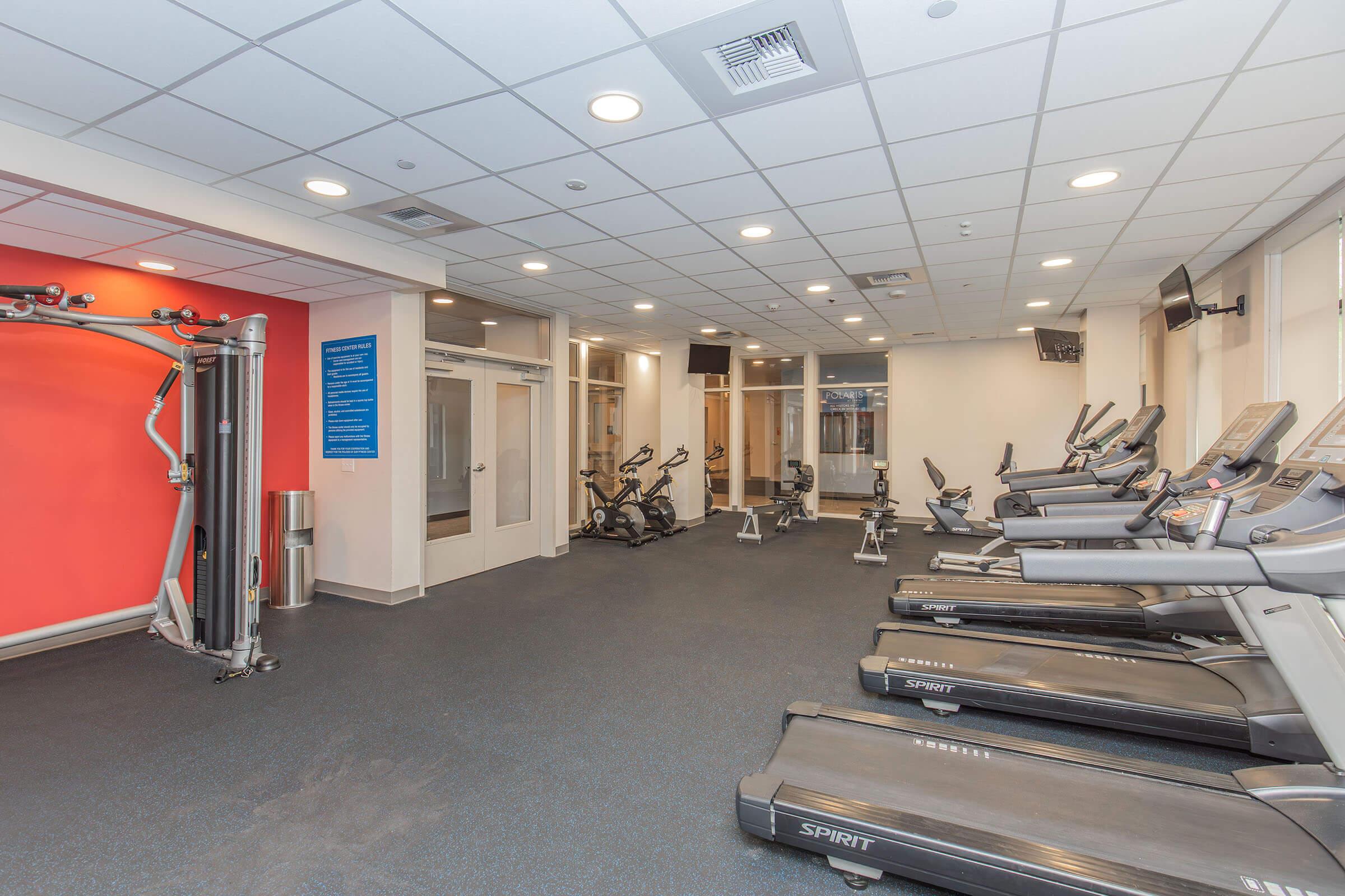 A spacious gym featuring various exercise equipment, including treadmills and stationary bikes. The room has bright, modern decor with an orange accent wall, and large windows that provide natural light. The floor is carpeted with black material, creating a welcoming workout environment.