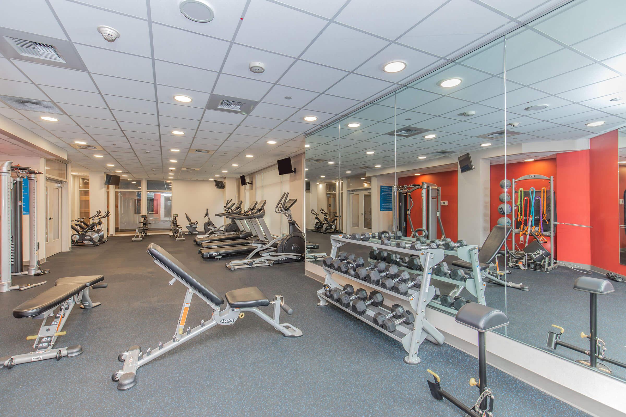 A modern fitness gym featuring workout equipment including treadmills, stationary bikes, and a weight bench. Dumbbell racks are visible along one wall, and large mirrors reflect the space, which has a clean and bright atmosphere. The room is well-lit with a minimalist design.