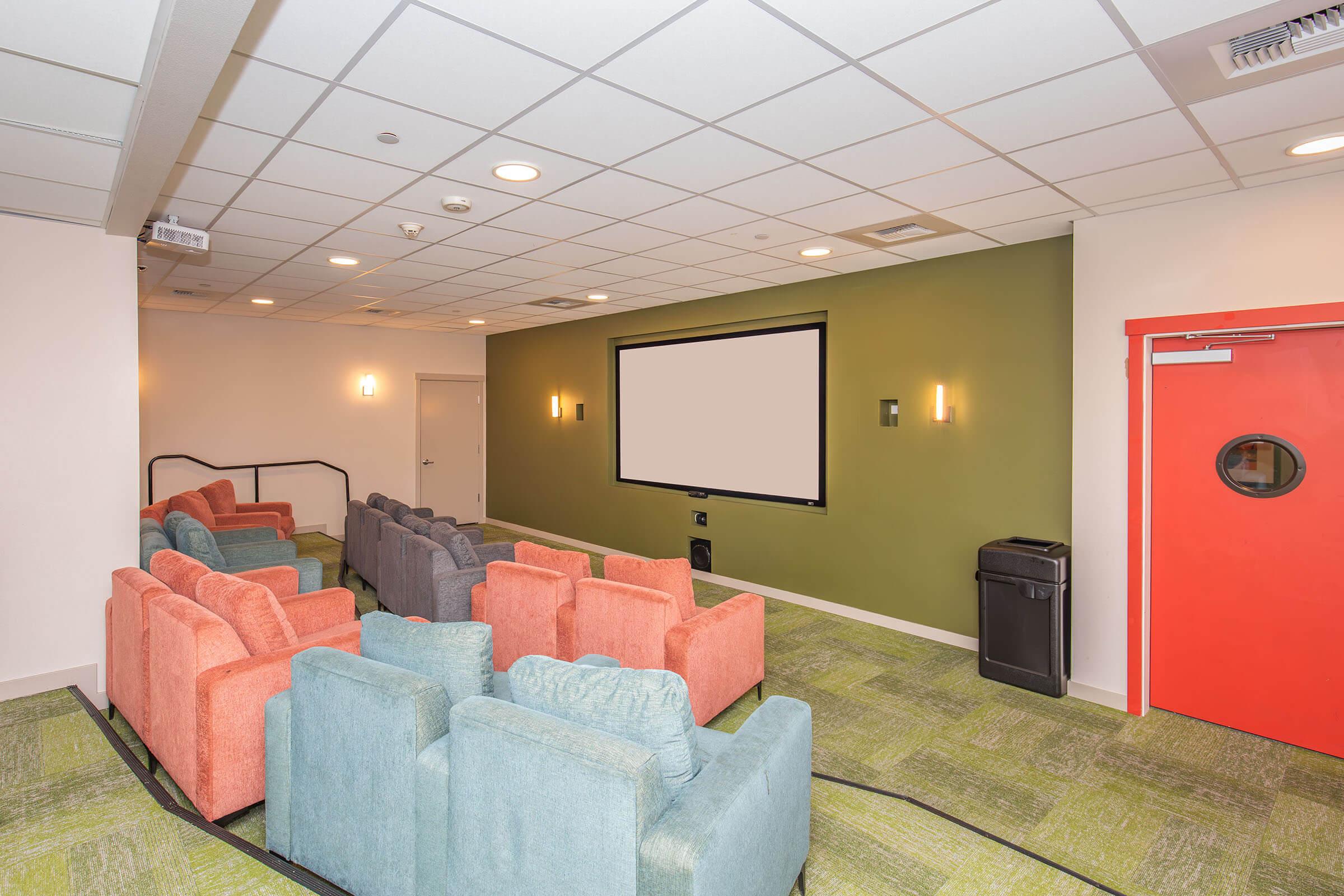 A cozy home theater room featuring colorful seating, including orange and teal armchairs, facing a large screen on a green accent wall. The room has soft lighting, a projector, and a red door, creating a welcoming atmosphere for watching movies or presentations.