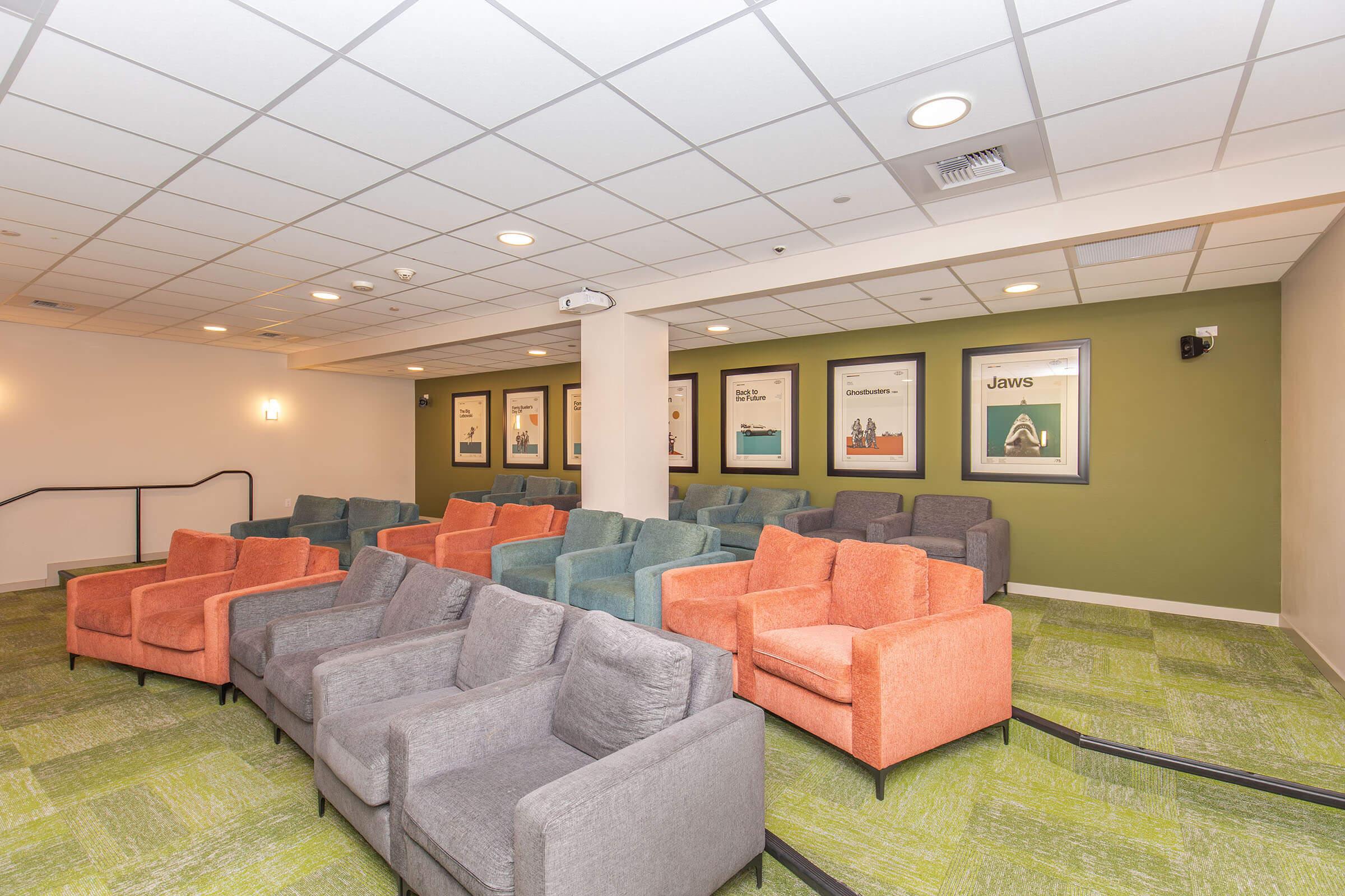A cozy lounge area featuring several colorful armchairs in grey, blue, and orange. The chairs are arranged in rows facing a blank wall, which has framed movie posters. The floor is covered with green carpeting, and the lighting is soft, creating a welcoming atmosphere for relaxation or viewing.