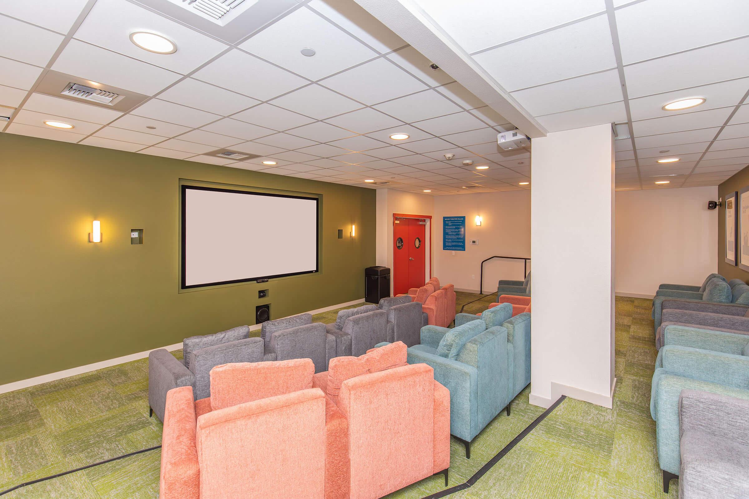 A cozy movie room with a large blank screen at the front. The space features several rows of colorful upholstered chairs in shades of orange, blue, and gray, arranged for group viewing. The walls are a mix of muted colors, and there are wall-mounted lights and a waste bin visible.