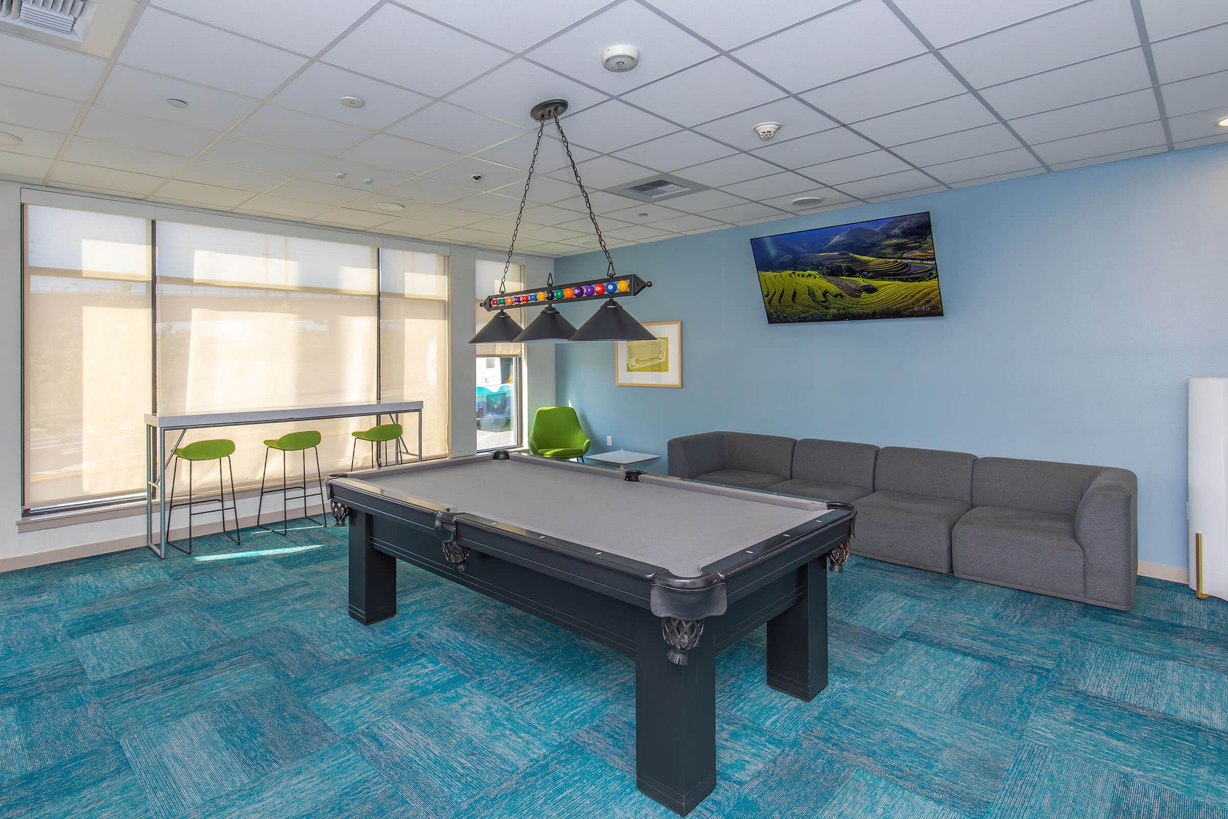A modern recreational room featuring a black pool table, green seating, and a cozy gray sofa. The walls are painted in soft blue tones, with large windows allowing natural light. A colorful pendant light hangs above the pool table, and a TV mounted on the wall displays a scenic landscape.