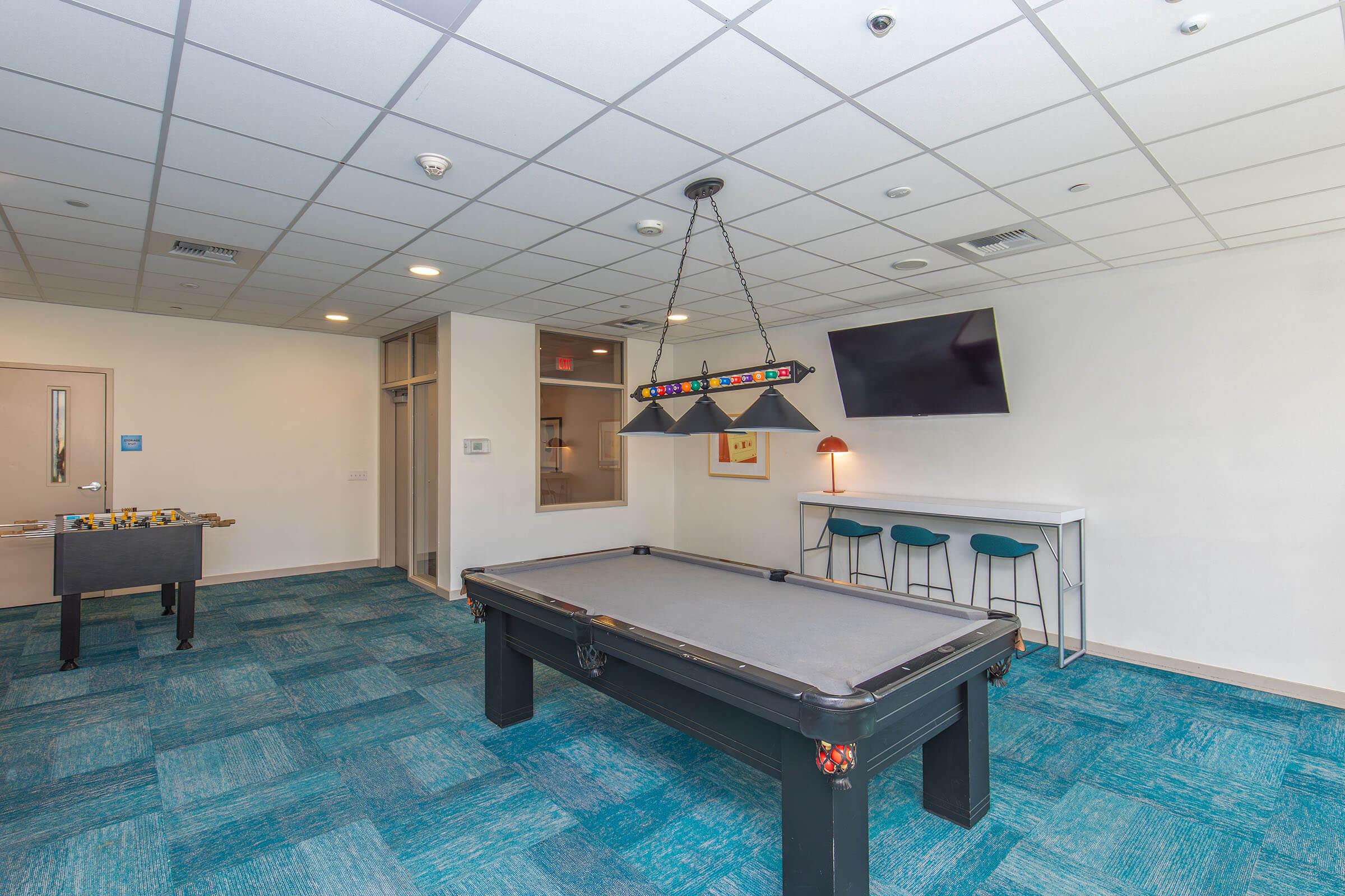 A well-lit recreational room featuring a pool table at the center, a foosball table on the left, and a small seating area with bar stools. A flat-screen TV is mounted on the wall, and there is a lamp on a small table. The floor is covered with blue carpeting, creating a modern and inviting space.