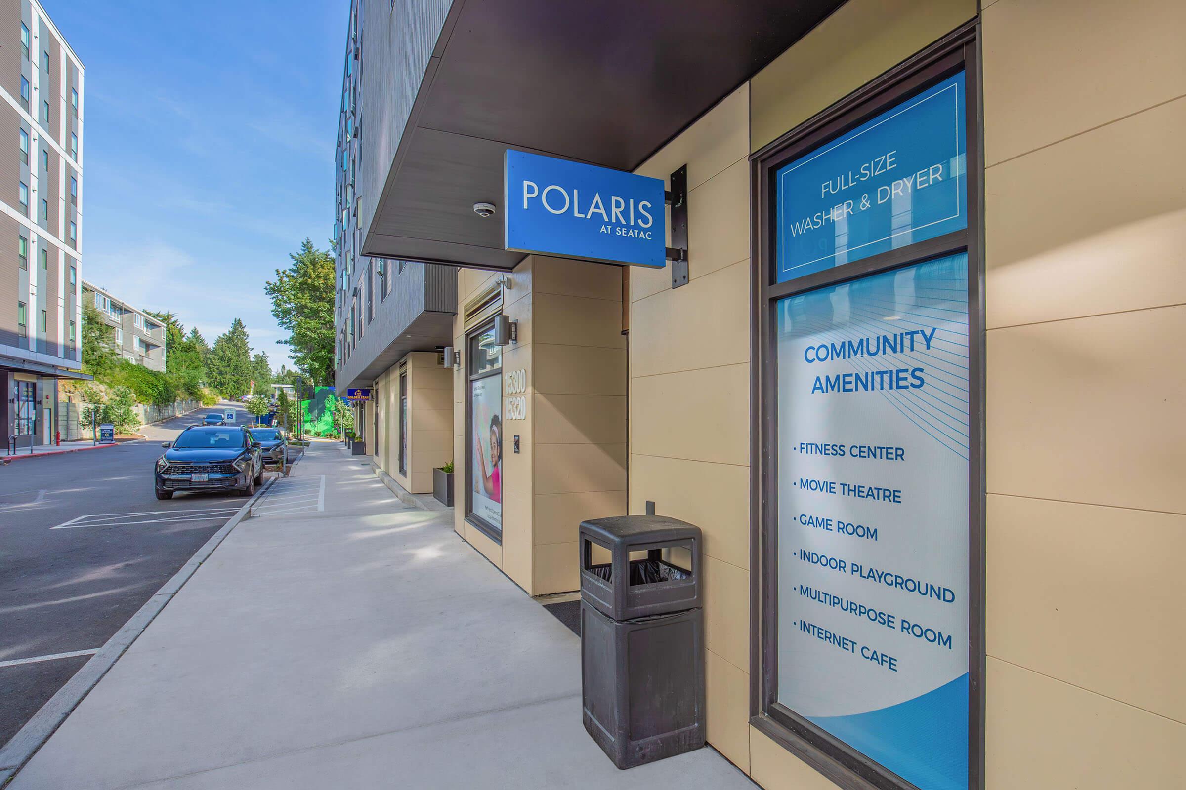 Signage for "Polaris at Beacon" showing community amenities such as a fitness center, movie theater, game room, indoor playground, multipurpose room, and internet cafe. The entrance is visible, along with a parking area and a trash can on the sidewalk.