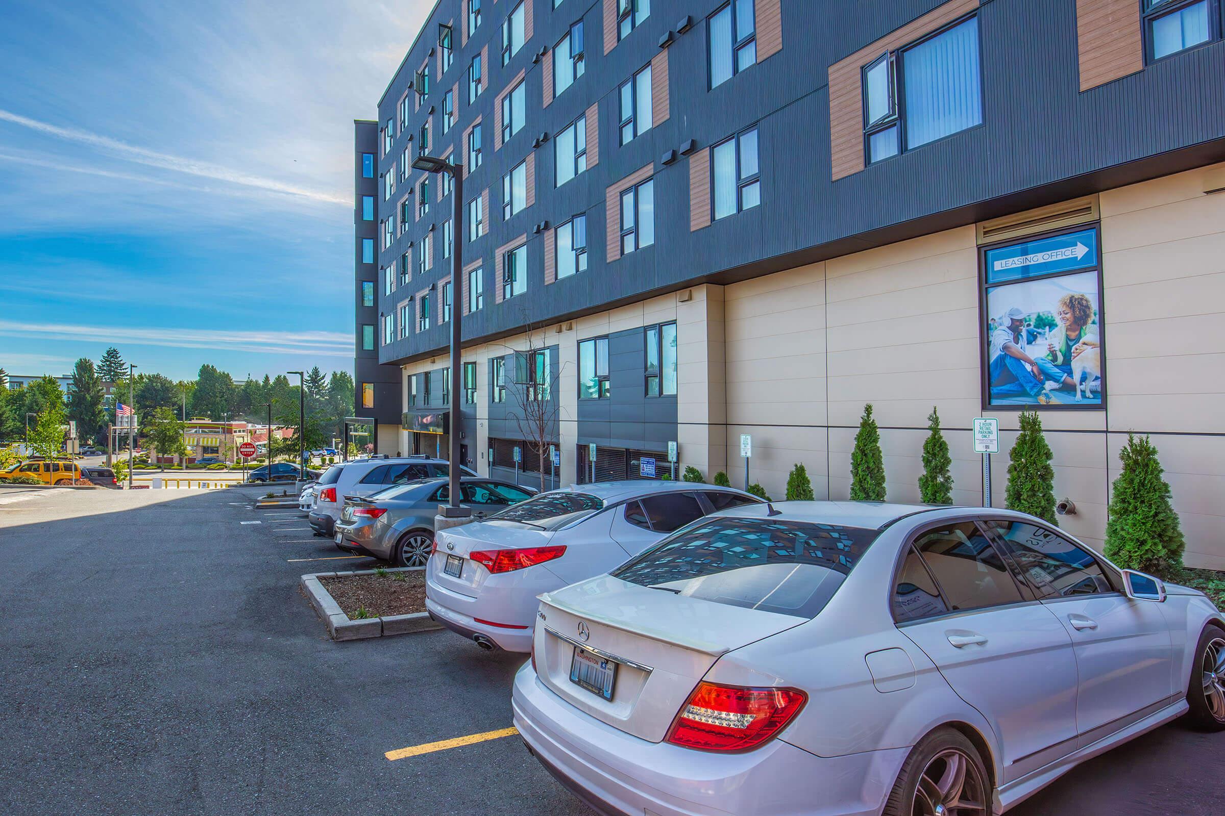 A parking lot with several cars parked in spaces, located next to a modern building featuring large windows and a promotional poster on the wall. The scene is set under a clear blue sky with some greenery in the background.