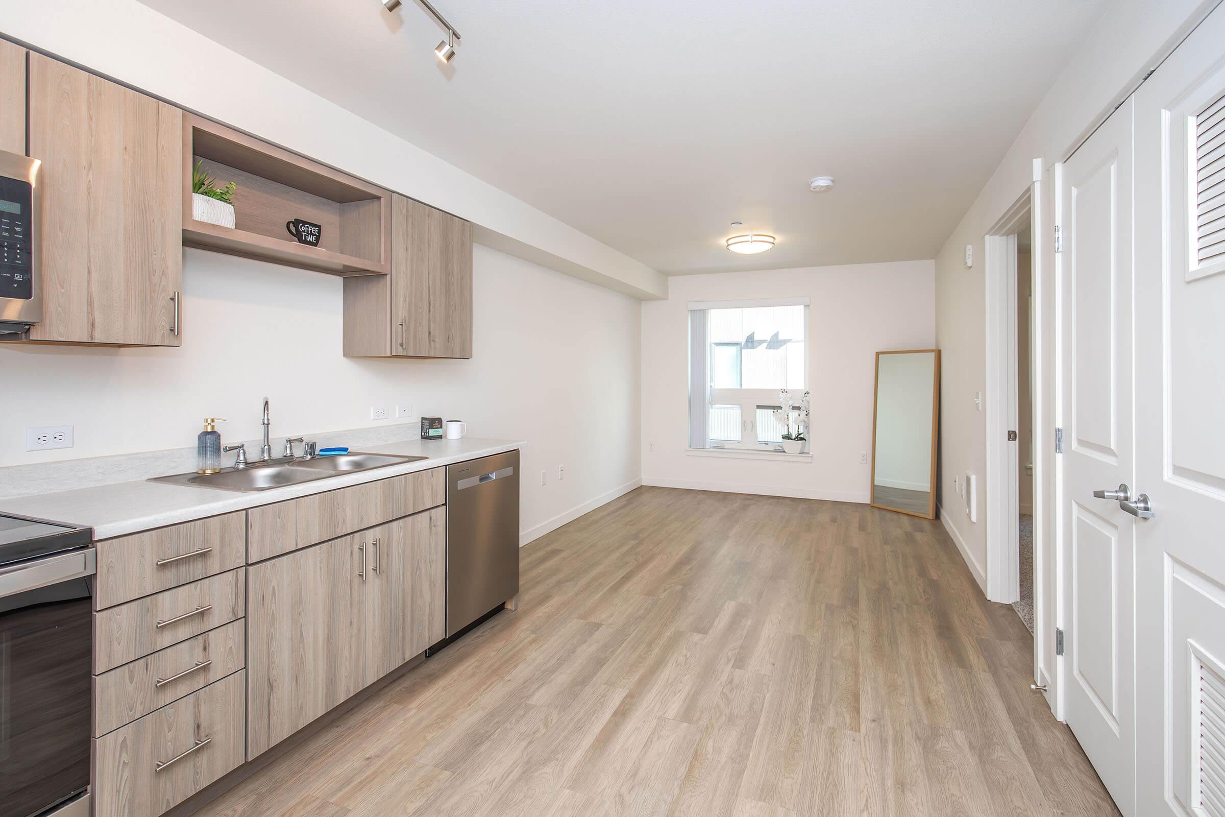 Modern kitchen and living area in an apartment. The kitchen features wooden cabinets, a stainless steel sink, and appliances. A mirror and a window are visible in the bright, empty space. The flooring is light-colored wood, creating a spacious, inviting atmosphere.
