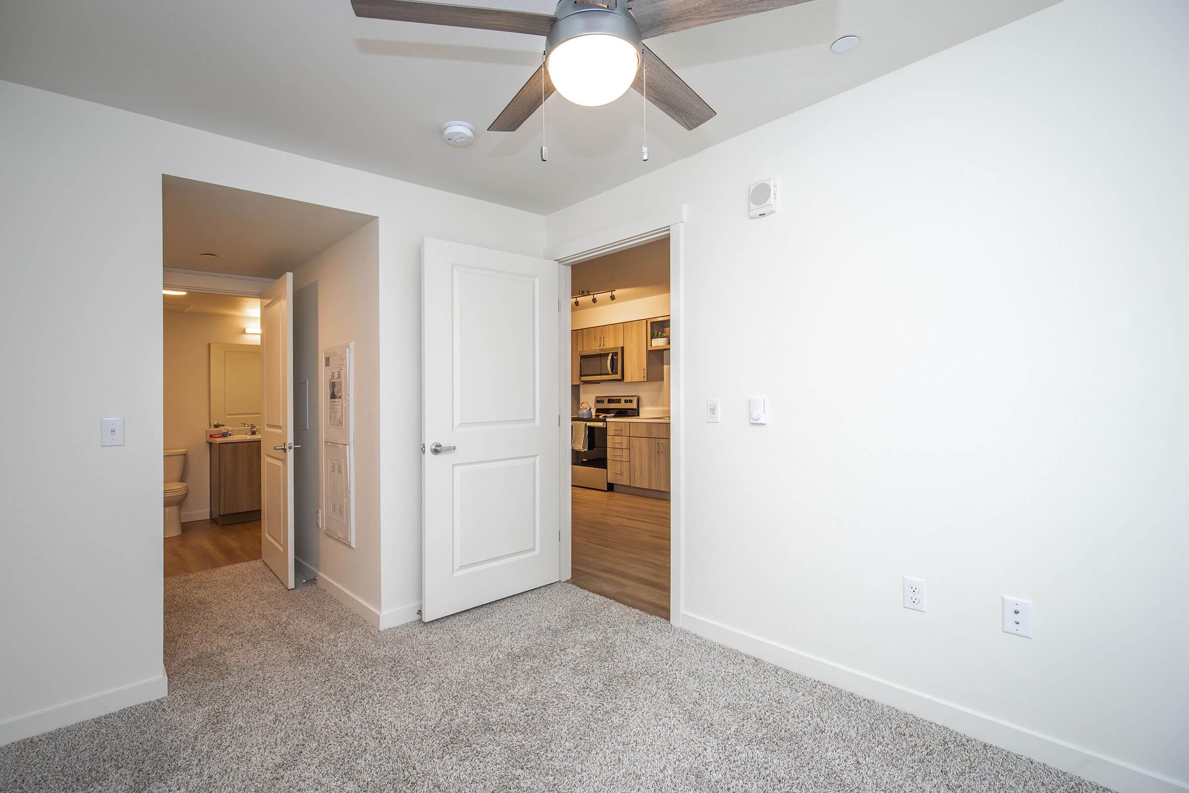 A well-lit, carpeted room with a ceiling fan, looking toward an open doorway that leads to a hallway and a bathroom. The opposite wall features another doorway leading to a kitchen area. The walls are painted in a light color, and there is a door partially visible in the background.