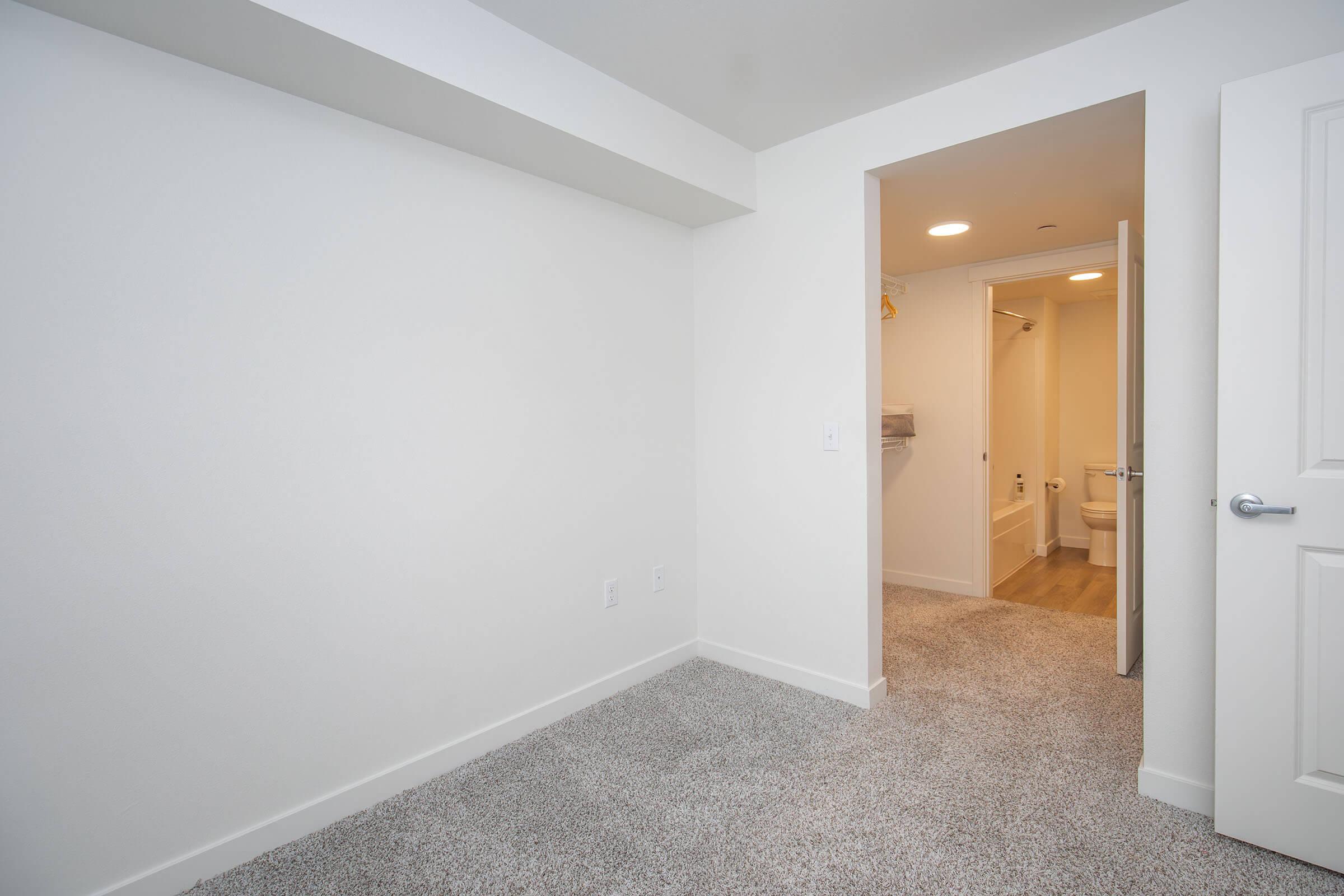 A spacious, empty room with light gray carpet and white painted walls. A doorway in the back leads to a bathroom area. The room is well-lit with natural light, giving a clean and modern feel. The absence of furniture emphasizes the room's size and potential for customization.
