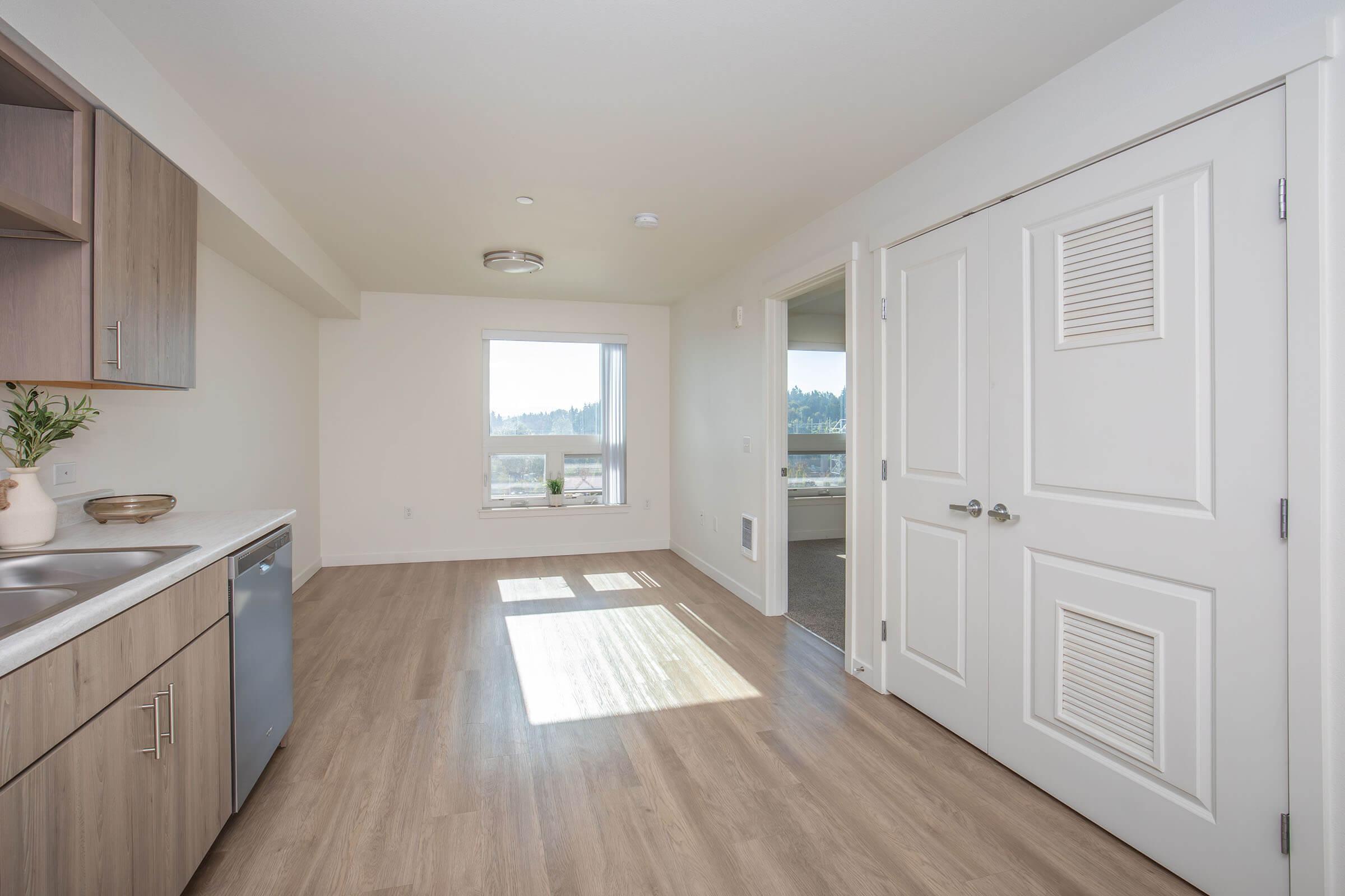 Modern, open-concept kitchen and living area with light-colored wood flooring. The kitchen includes cabinetry and a stainless steel dishwasher. A large window lets in natural light, and the space features neutral-colored walls. A doorway leads to another room.