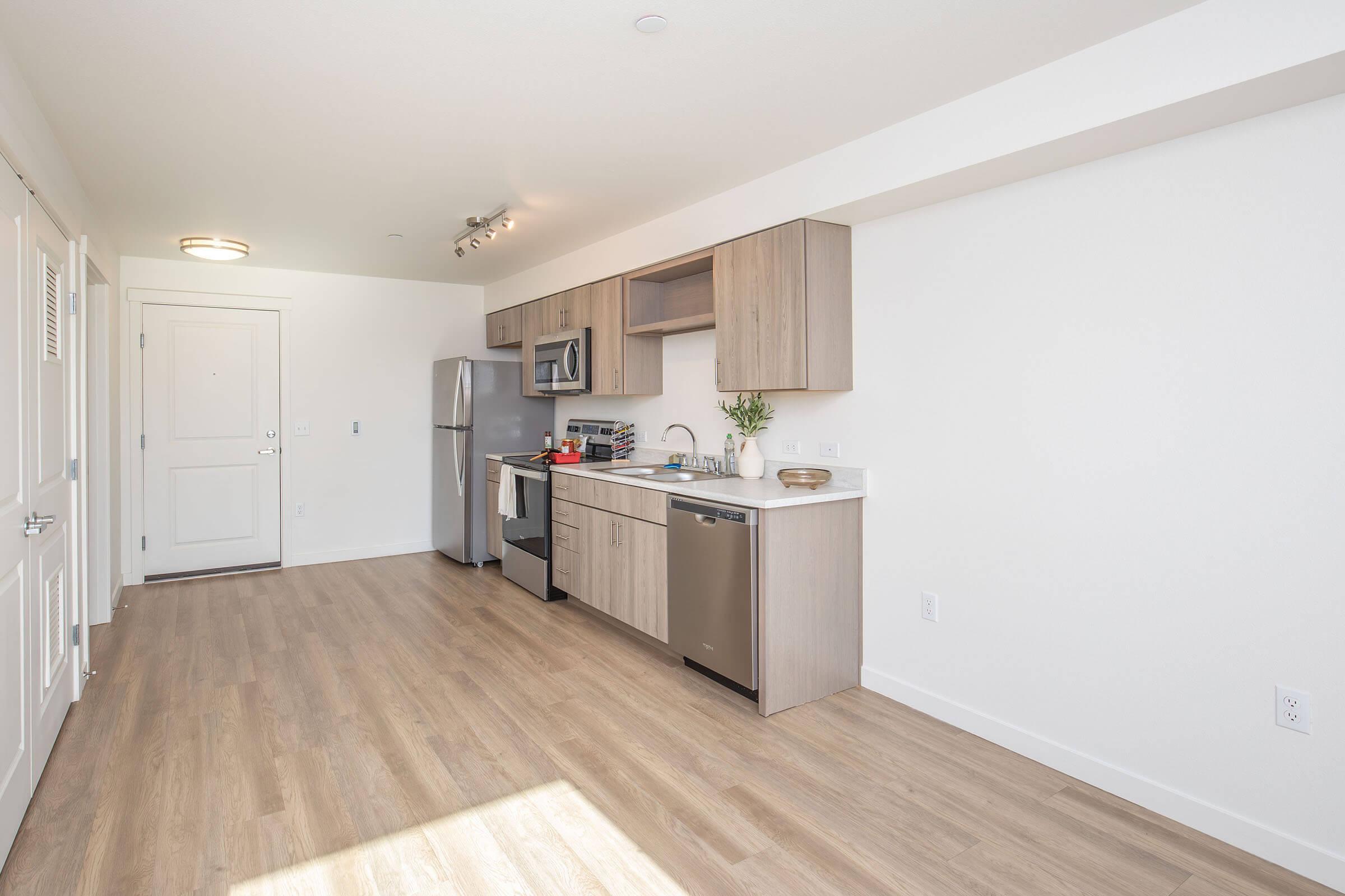 Modern kitchen with light wood cabinetry, stainless steel appliances including a refrigerator, dishwasher, and oven. The space features a contemporary design with white walls and wood flooring, a minimalist aesthetic, and a small dining area. Natural light fills the room through a nearby door.