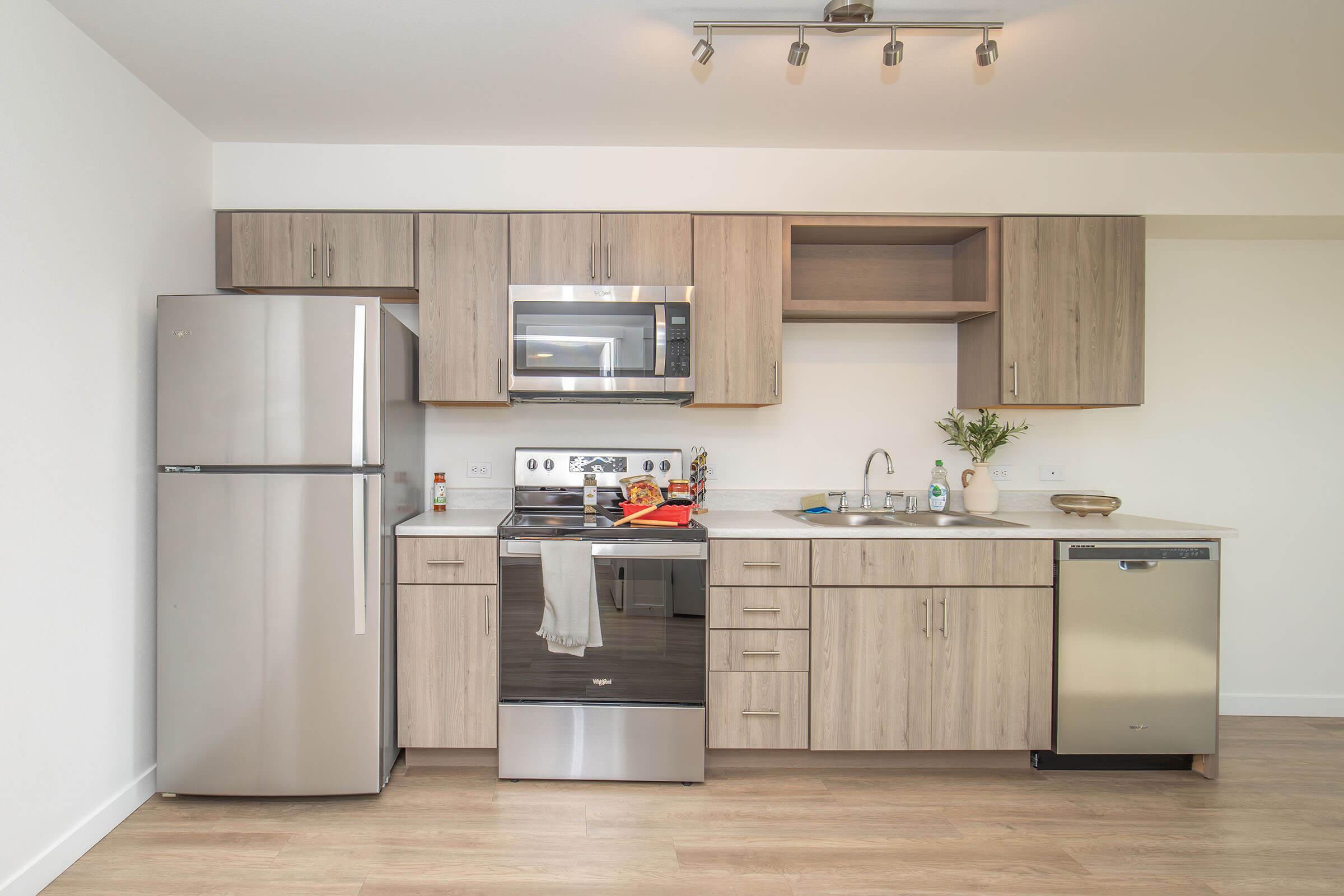 Modern kitchen featuring stainless steel appliances, including a refrigerator, microwave, and oven. The cabinetry is light wood, with an open countertop area and sink. A small vase with greenery is placed on the counter, and there are kitchen utensils and items on the surface. Bright, well-lit space with a clean design.