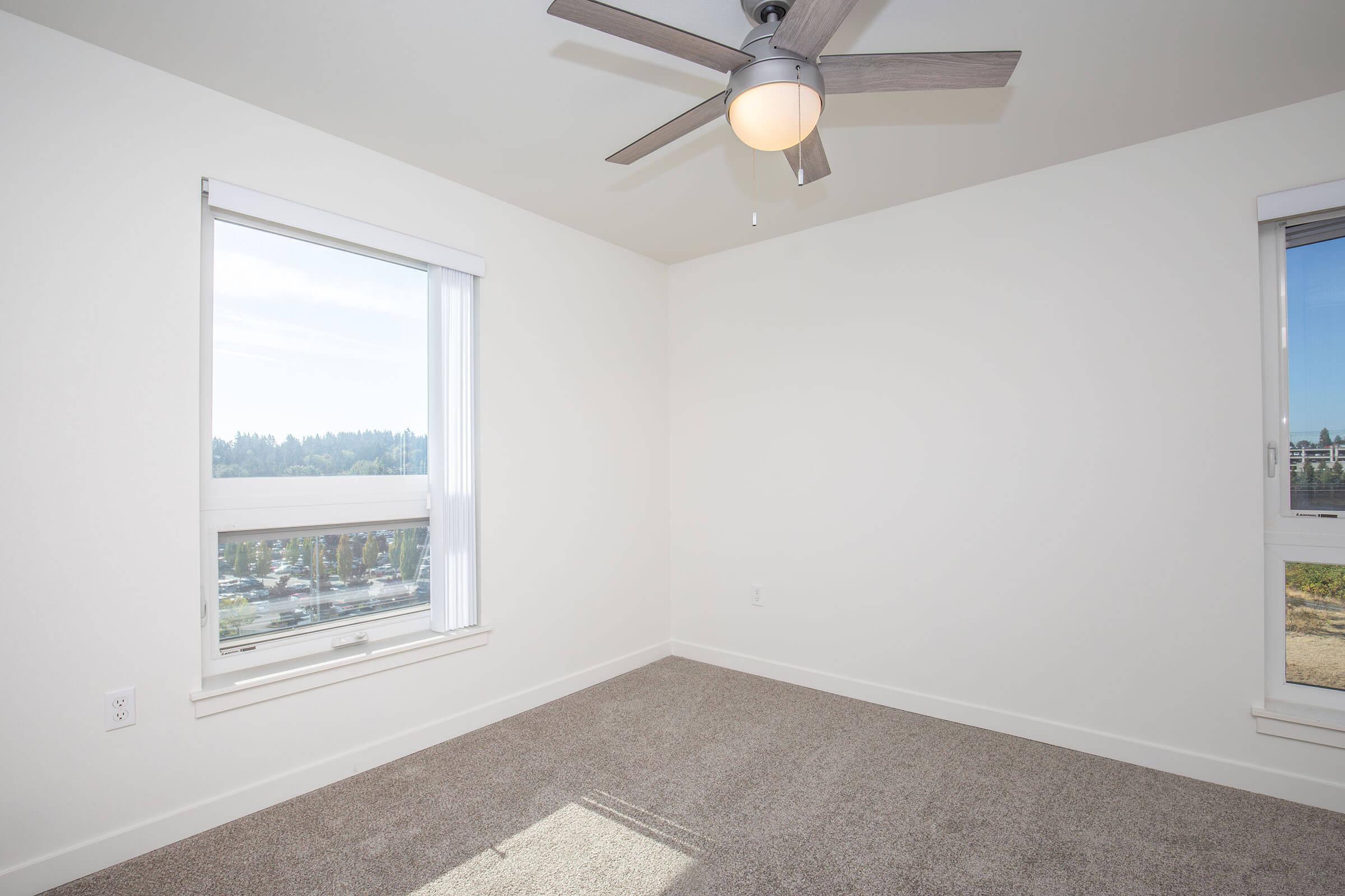 A bright, empty room with light-colored walls and a light gray carpet. It features a ceiling fan and two windows with white blinds, offering a view of greenery outside. The space is well-lit, creating a welcoming atmosphere, ideal for various uses.