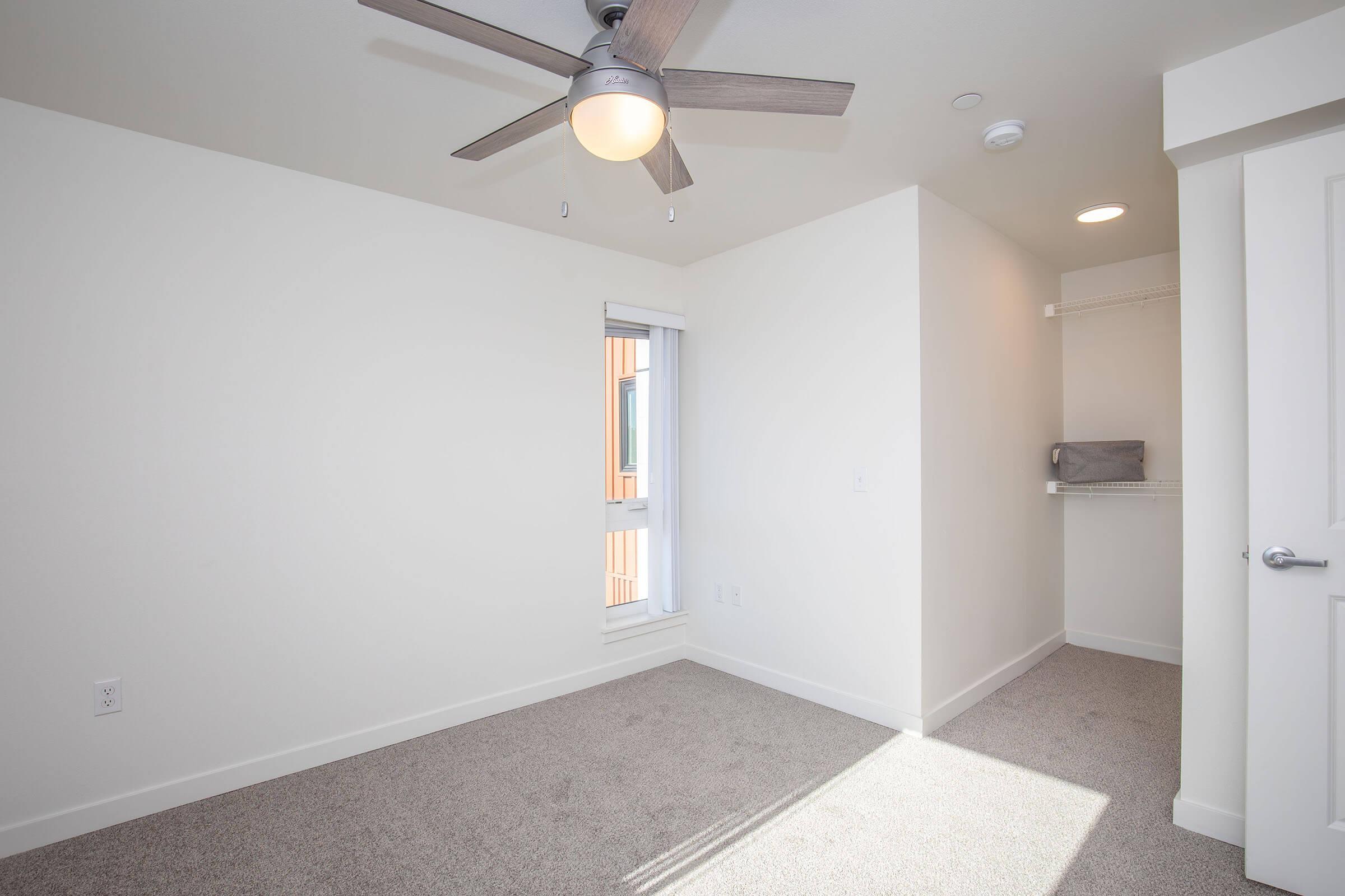 A bright, empty room with light-colored walls and carpet. It features a ceiling fan and a window letting in natural light. A small, built-in shelf is visible in the corner, along with a closet space and modern overhead lighting.