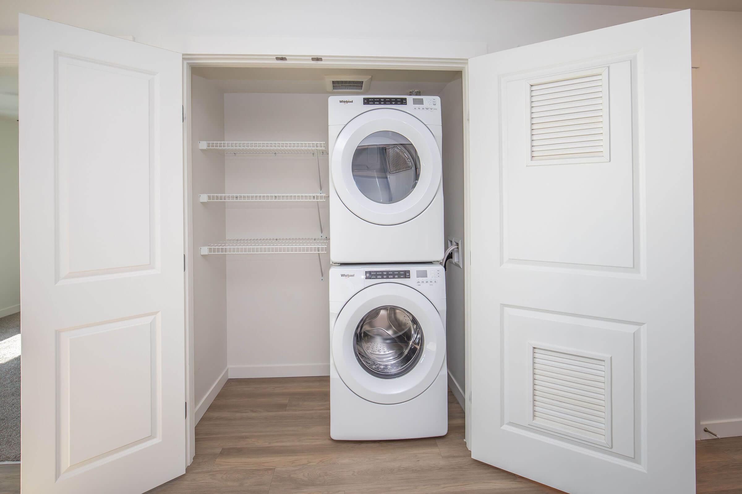 A laundry closet featuring a stacked washer and dryer in white. The doors are open, revealing the appliances and some shelves for storage. The interior is well-lit and has a neutral color scheme with wooden flooring.