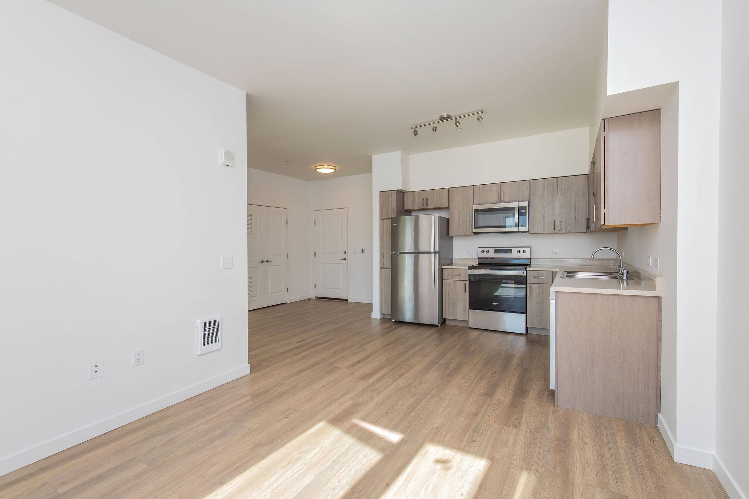 Interior view of a modern, open-concept kitchen and living area. The kitchen features stainless steel appliances, including a refrigerator and oven, with wooden cabinets and a light-colored countertop. The flooring is hardwood, and the walls are painted white, providing a bright, spacious atmosphere.