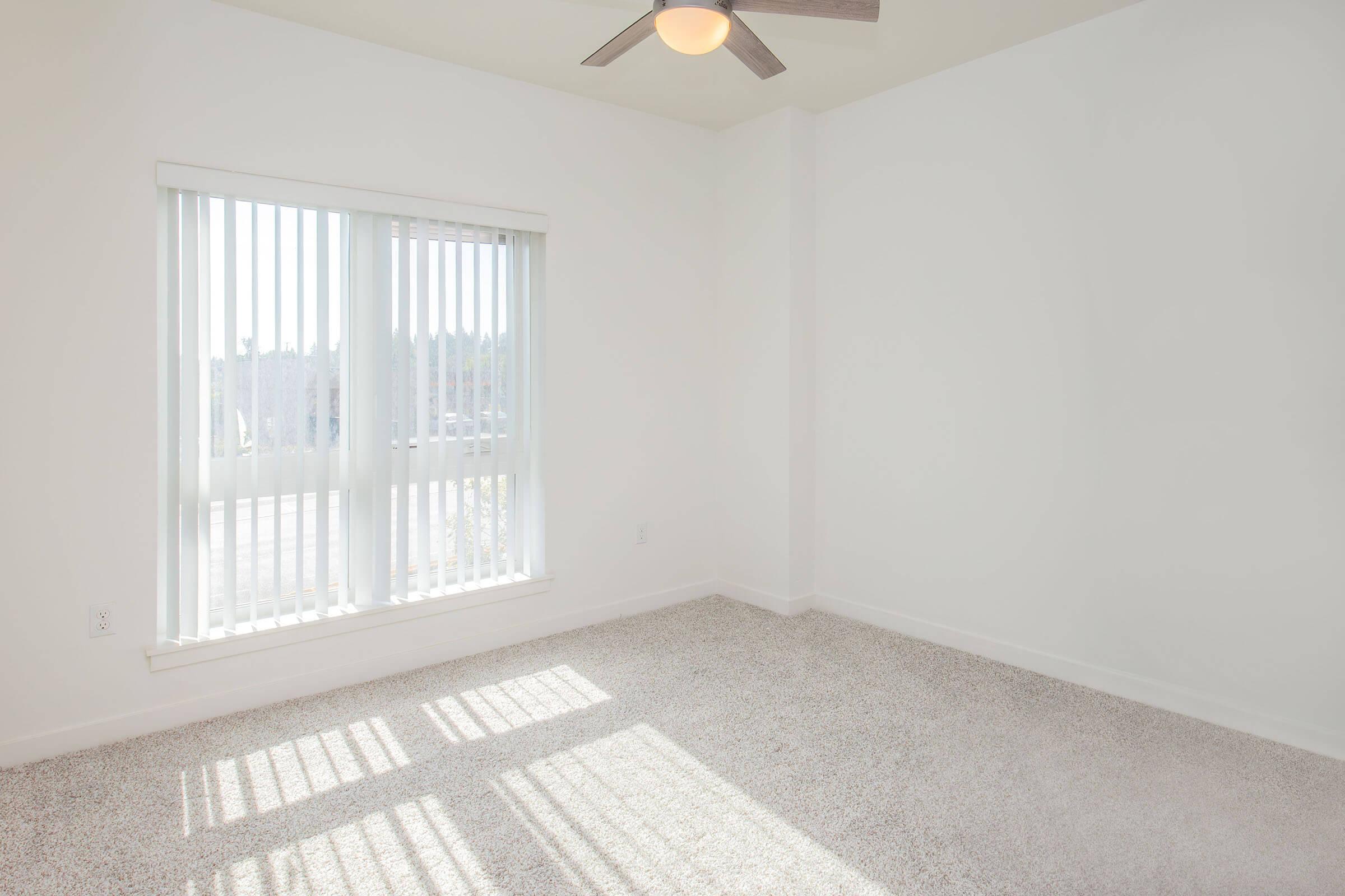 A bright, sparsely furnished room with beige carpet and white walls. It features large vertical blinds covering a window that lets in natural light. A ceiling fan is visible, and the space appears clean and minimalistic, suitable for a bedroom or office.