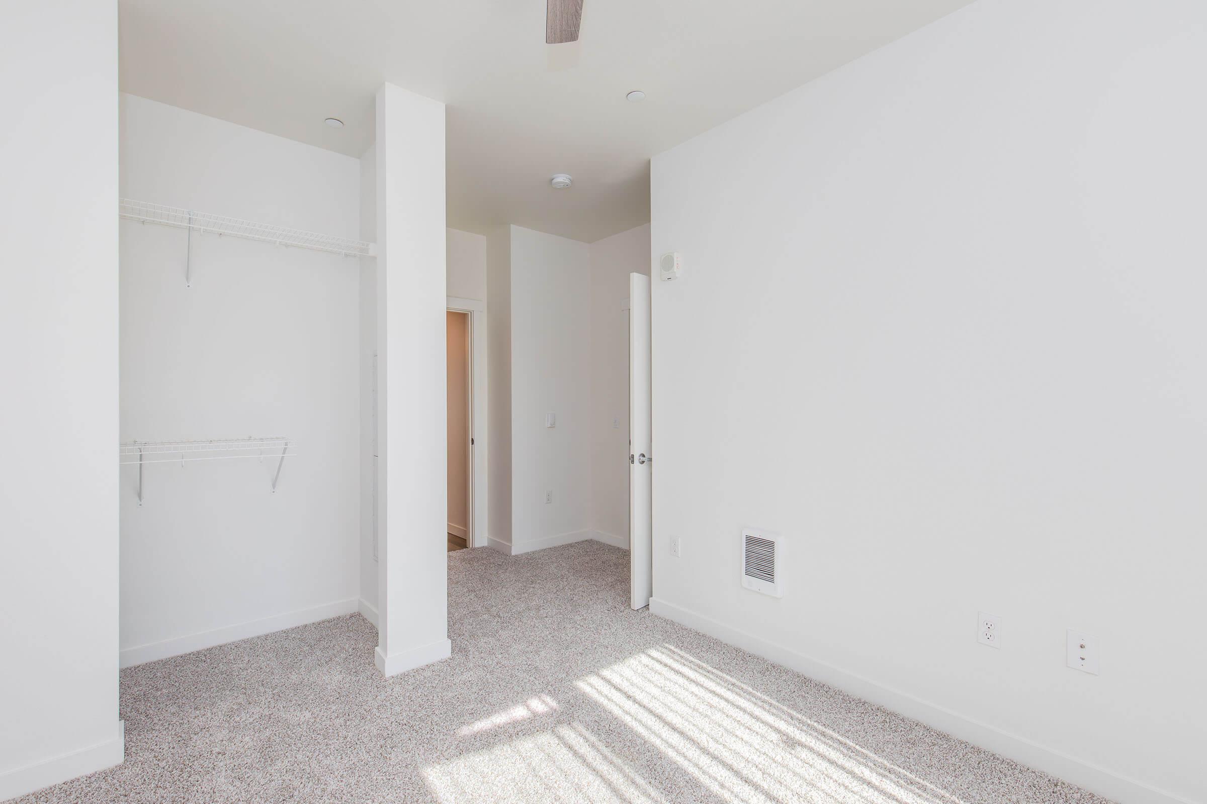A bright, empty room with light-colored walls and carpet. There is a closet with wire shelving on the left, a door leading to another room in the background, and a vent on the floor. The room has natural light streaming in, creating a clean and spacious atmosphere.