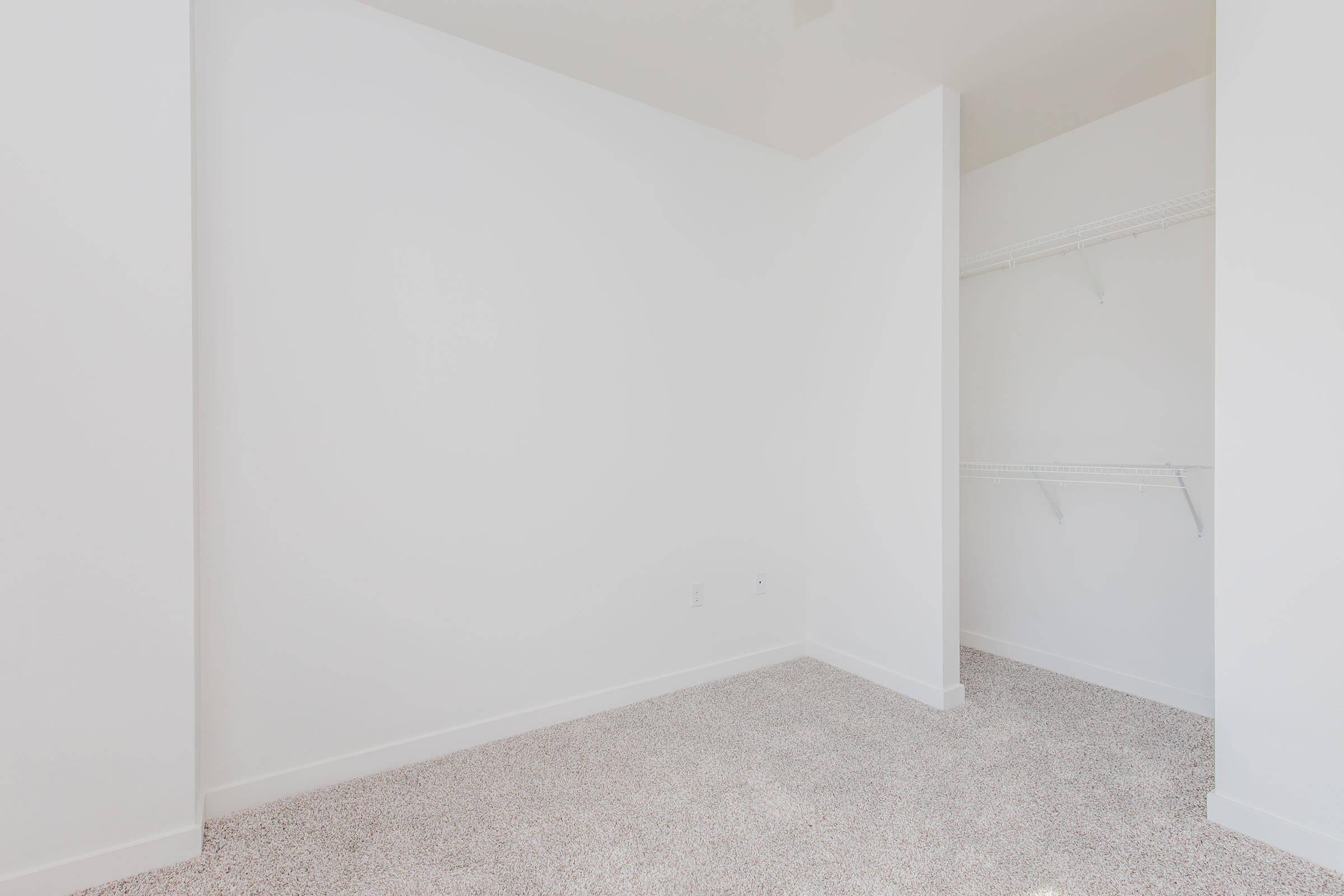 A bright, minimalist room with white walls and a light carpet. The space features a section of a closet with shelves on the left, offering clean lines and a simple design. The overall atmosphere is open and uncluttered, creating a sense of calm and spaciousness.