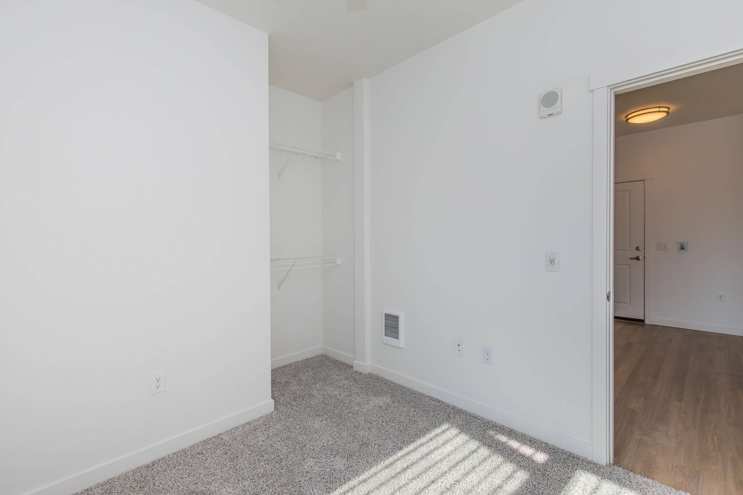A clean, empty room featuring light-colored walls and carpeted flooring. There are shelves mounted on one wall, and a doorway leading to another area. Natural light streams in through an unseen window, creating a bright atmosphere. The room is simple and uncluttered, suitable for various uses.