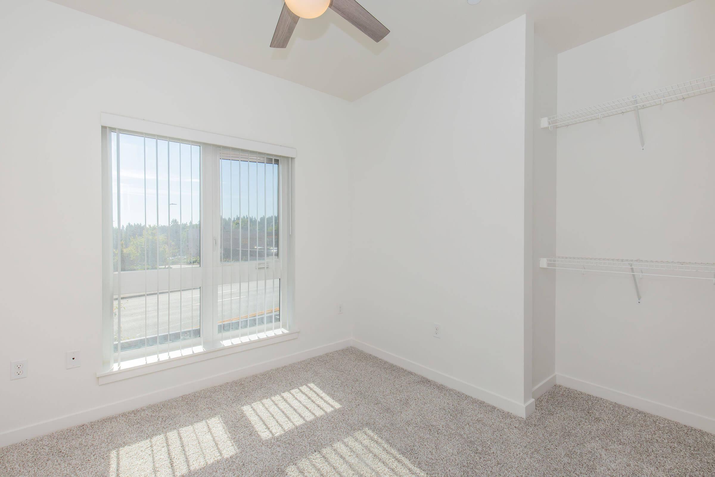 A bright, empty bedroom with a large window featuring blinds. Sunlight casts shadows on the carpeted floor. The room has a ceiling fan and a small closet on the right side with shelves. The walls are painted white, giving a clean and modern appearance.