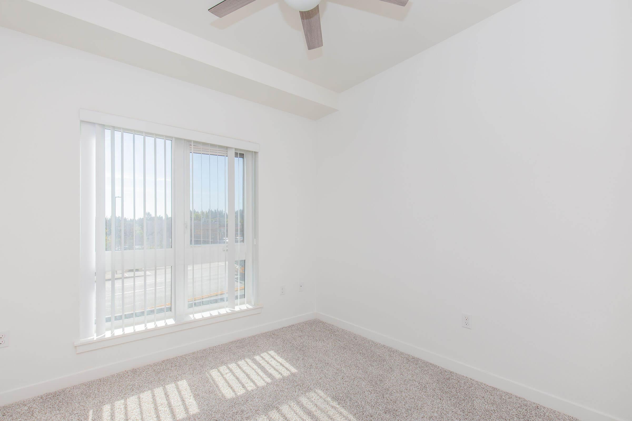 A bright, empty room featuring a white ceiling and light-colored walls. The floor is covered in soft beige carpet. A large window with horizontal blinds lets in natural light, casting shadows onto the floor. A ceiling fan is mounted above, adding to the airy feel of the space.