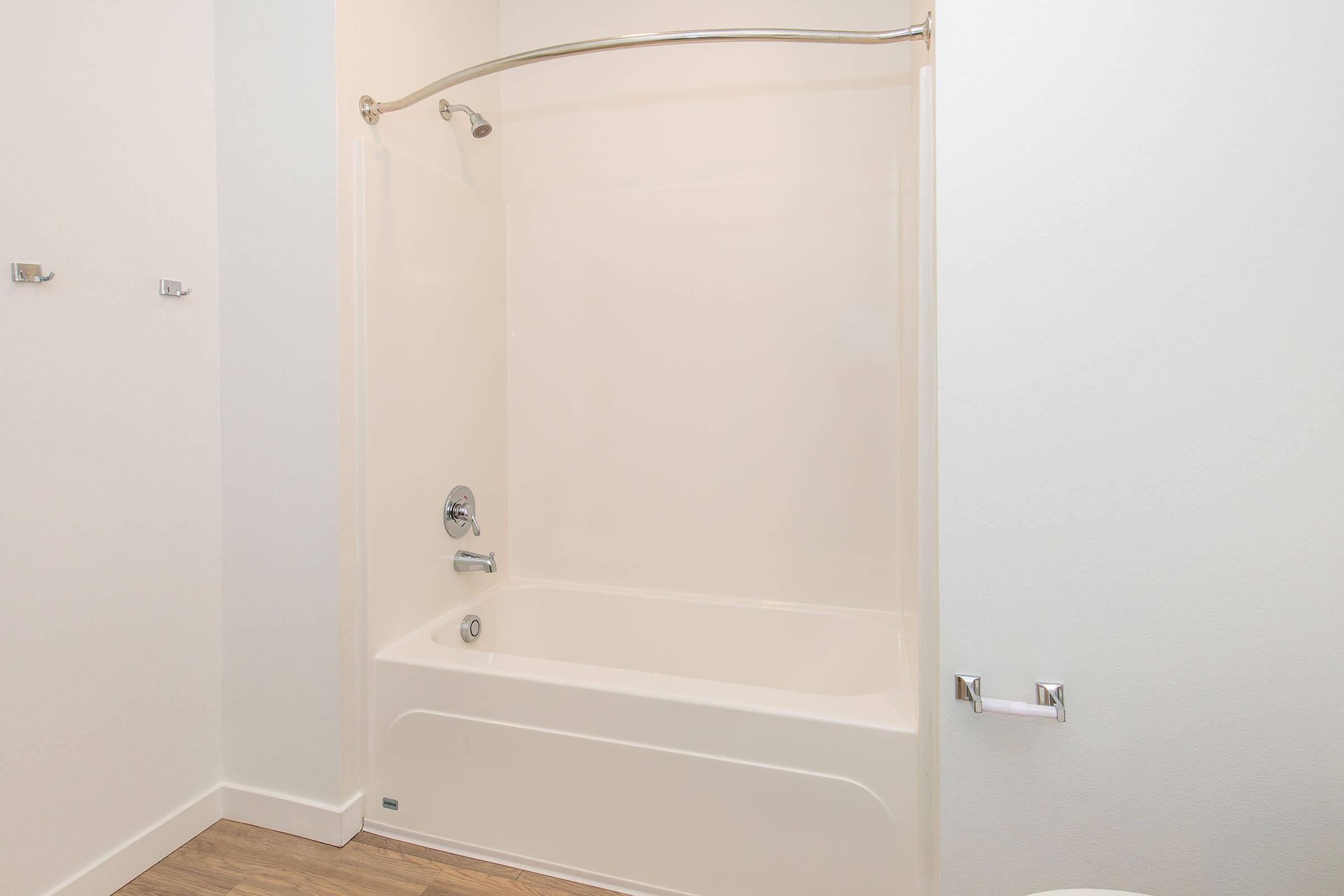 A clean, modern bathroom featuring a white tub with a shower curtain. There is a chrome faucet and showerhead, and the walls are plain white. The floor has light wood-like tiles. Hooks and a toilet paper holder are mounted on the wall, contributing to the minimalist design.