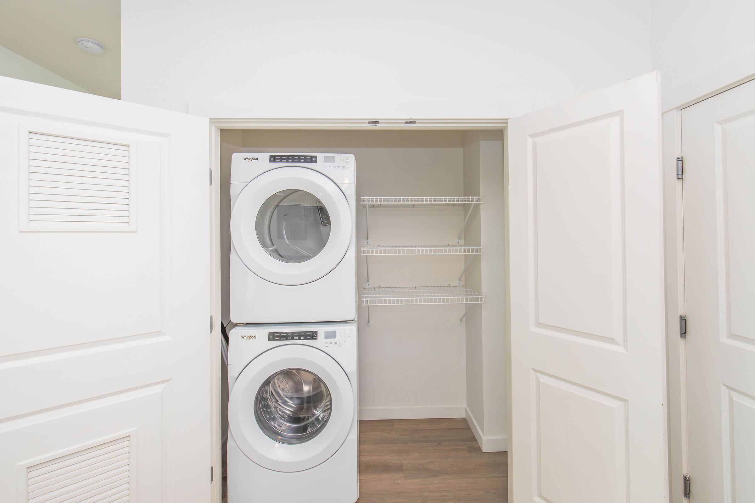 A laundry closet featuring a stacked washer and dryer, with white shelving on the side. The walls are painted white and the flooring is light wood. The closet doors are open, providing a clear view of the appliances and storage space.
