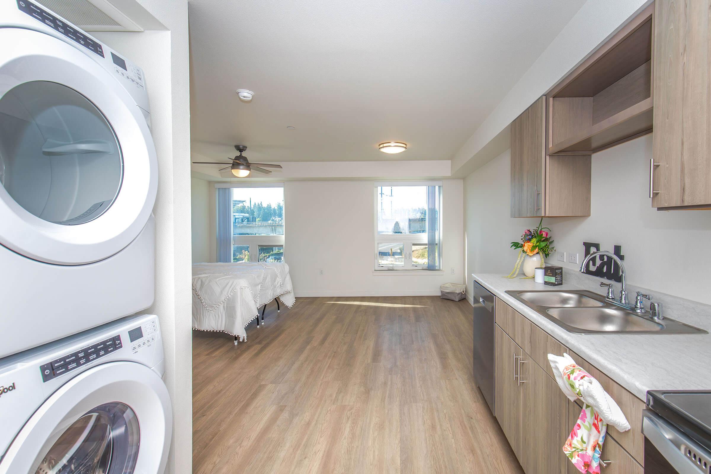 A modern apartment interior featuring a laundry area with stacked washer and dryer on the left, a kitchenette with dark wood cabinetry and stainless steel appliances, and a small living space with a ceiling fan and large windows. The room is bright with light-colored walls and wood flooring.