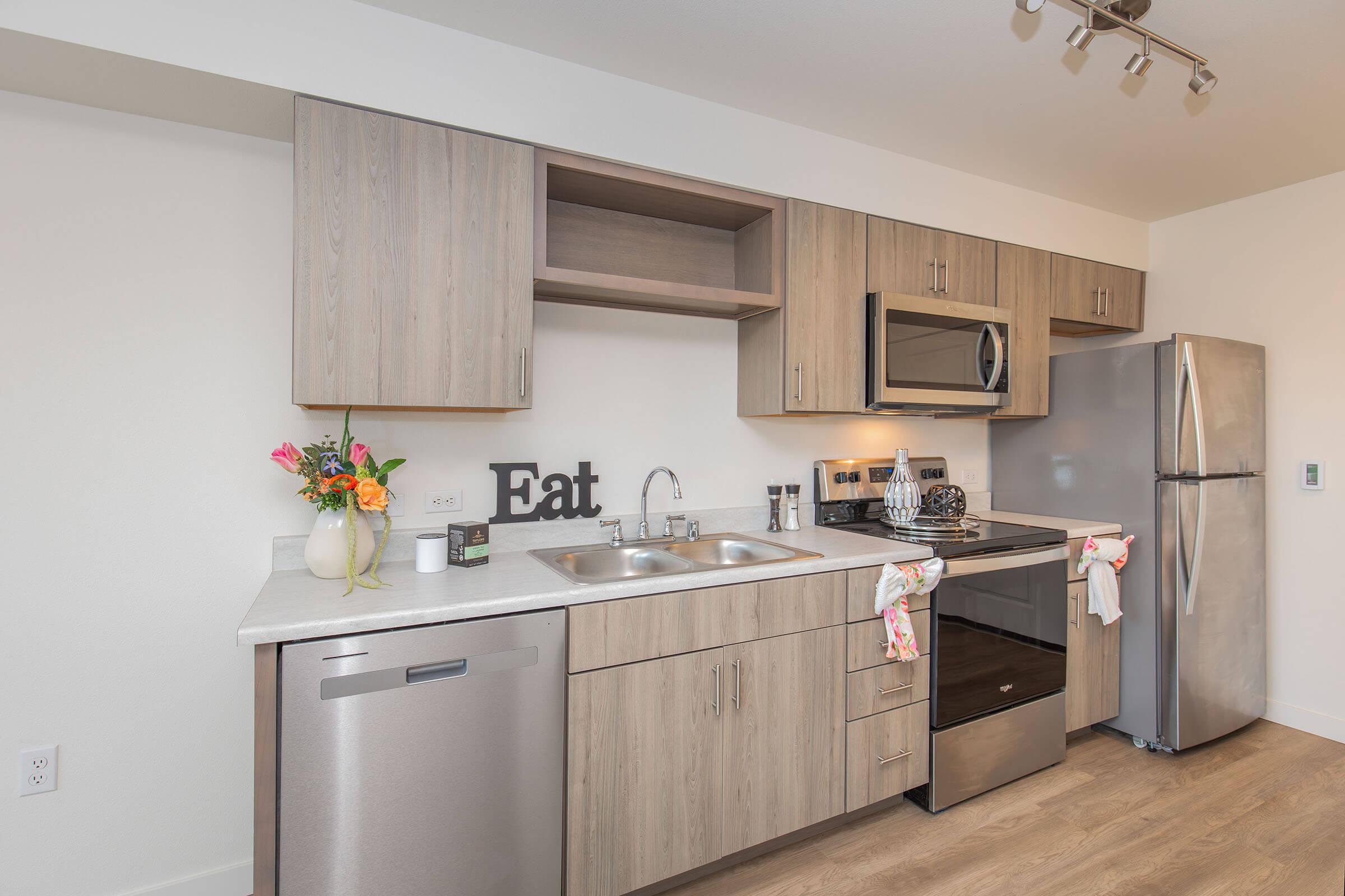 Modern kitchen featuring light wood cabinetry, stainless steel appliances including a refrigerator and microwave, and a double sink. A decorative "Eat" sign is displayed on the counter alongside a vase of flowers and kitchen utensils. Soft lighting enhances the contemporary design.