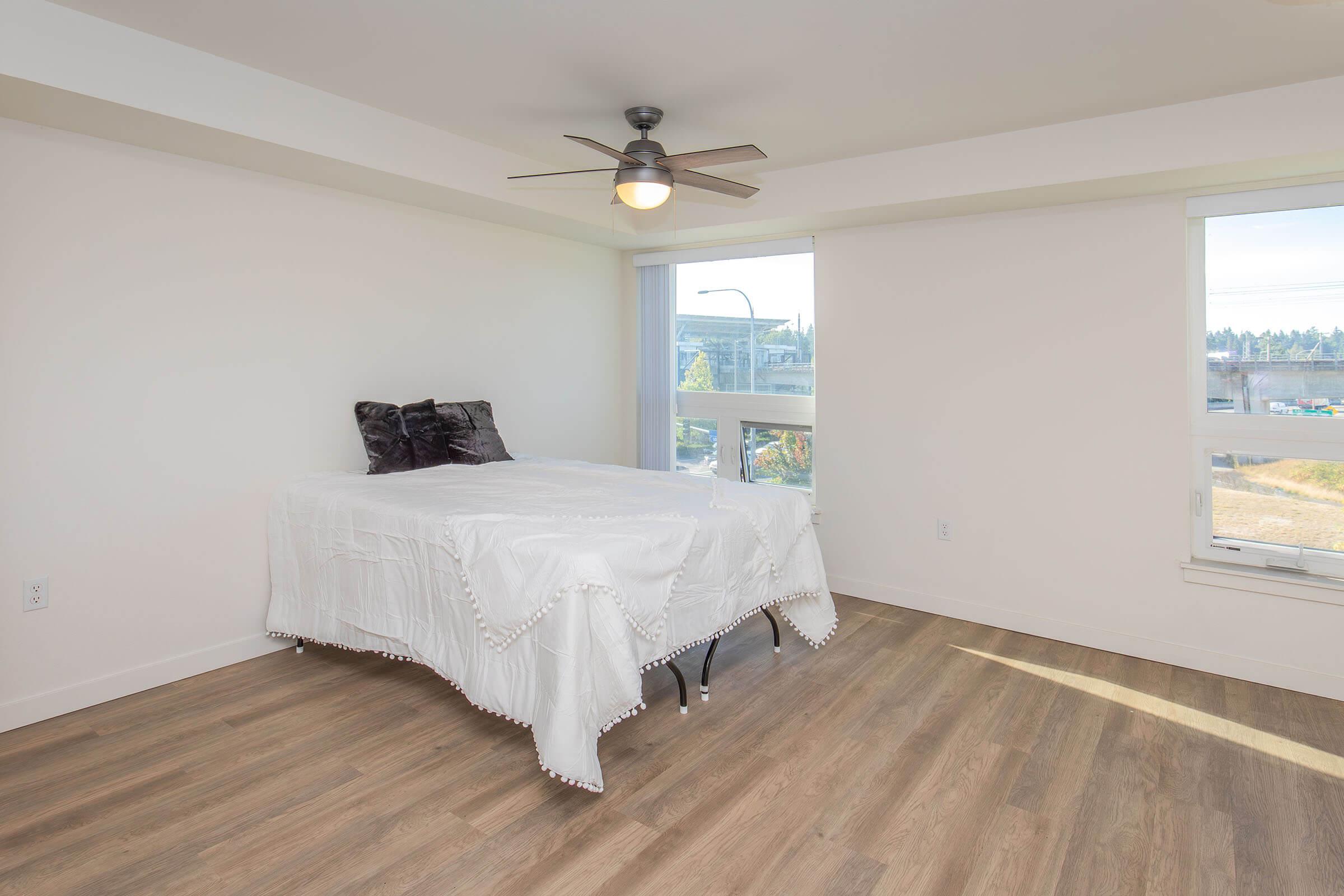 A spacious, minimalist bedroom featuring a bed with a white blanket, two dark pillows, and a ceiling fan. Natural light floods the room through large windows, revealing a view of an outdoor area. The floor is made of light-colored wood, enhancing the airy atmosphere.