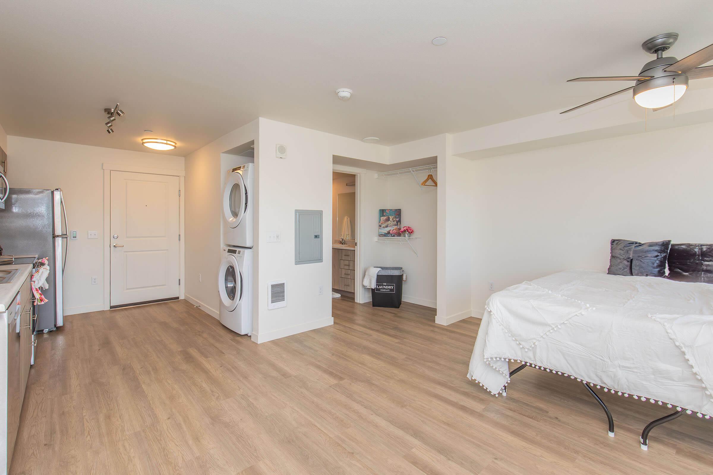 A modern studio apartment featuring an open layout with a kitchen, living space, and a bed. The space includes a ceiling fan, stainless steel appliances, a washer and dryer, and light wood flooring. The walls are painted white, creating a bright and airy atmosphere.