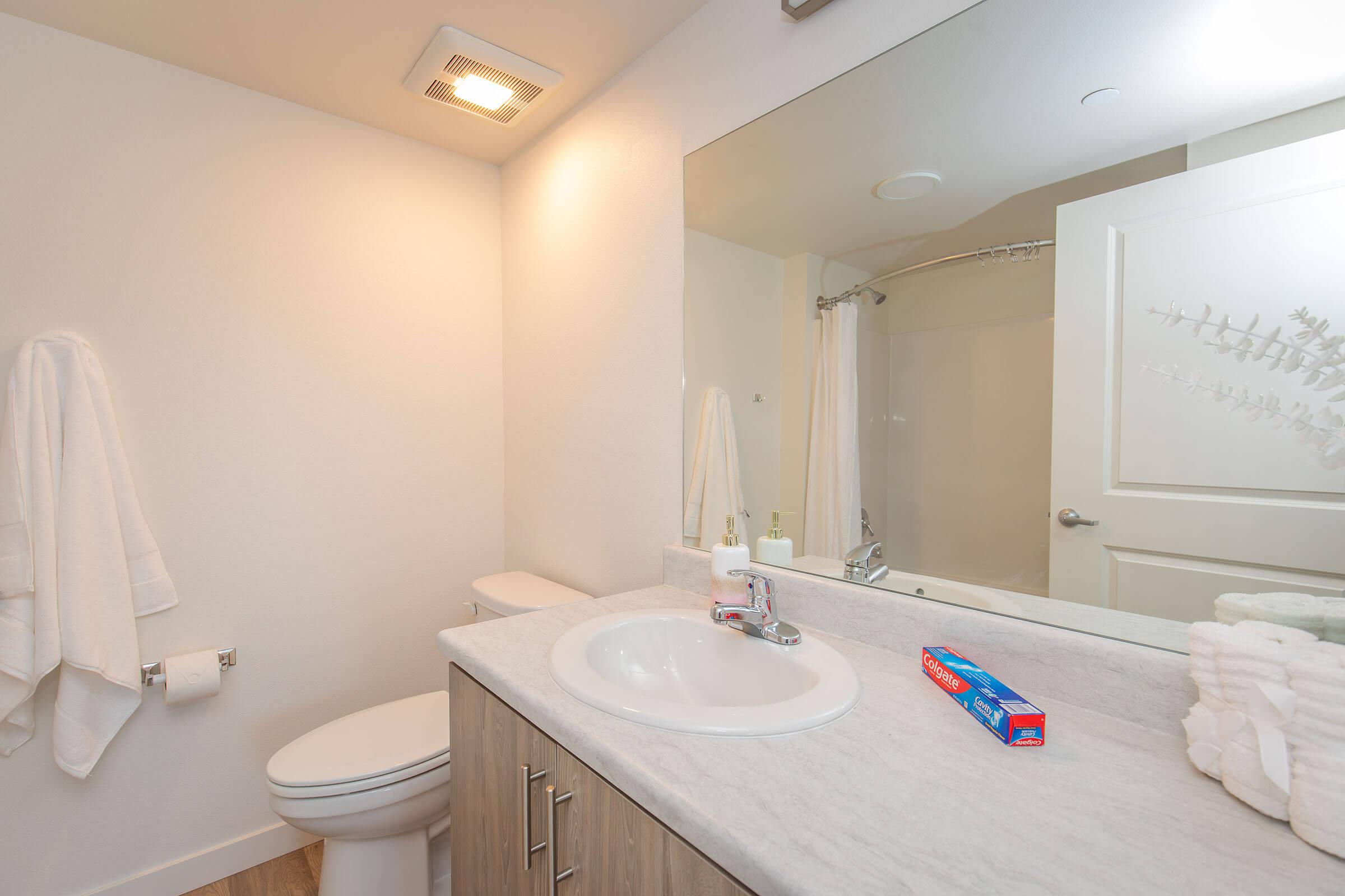 A modern bathroom featuring a white sink with a chrome faucet, a toilet, and a large mirror. There are neatly arranged towels hanging on the wall, a shower curtain in the background, and a tube of toothpaste on the counter. The decor is clean and minimalistic, with neutral tones throughout.