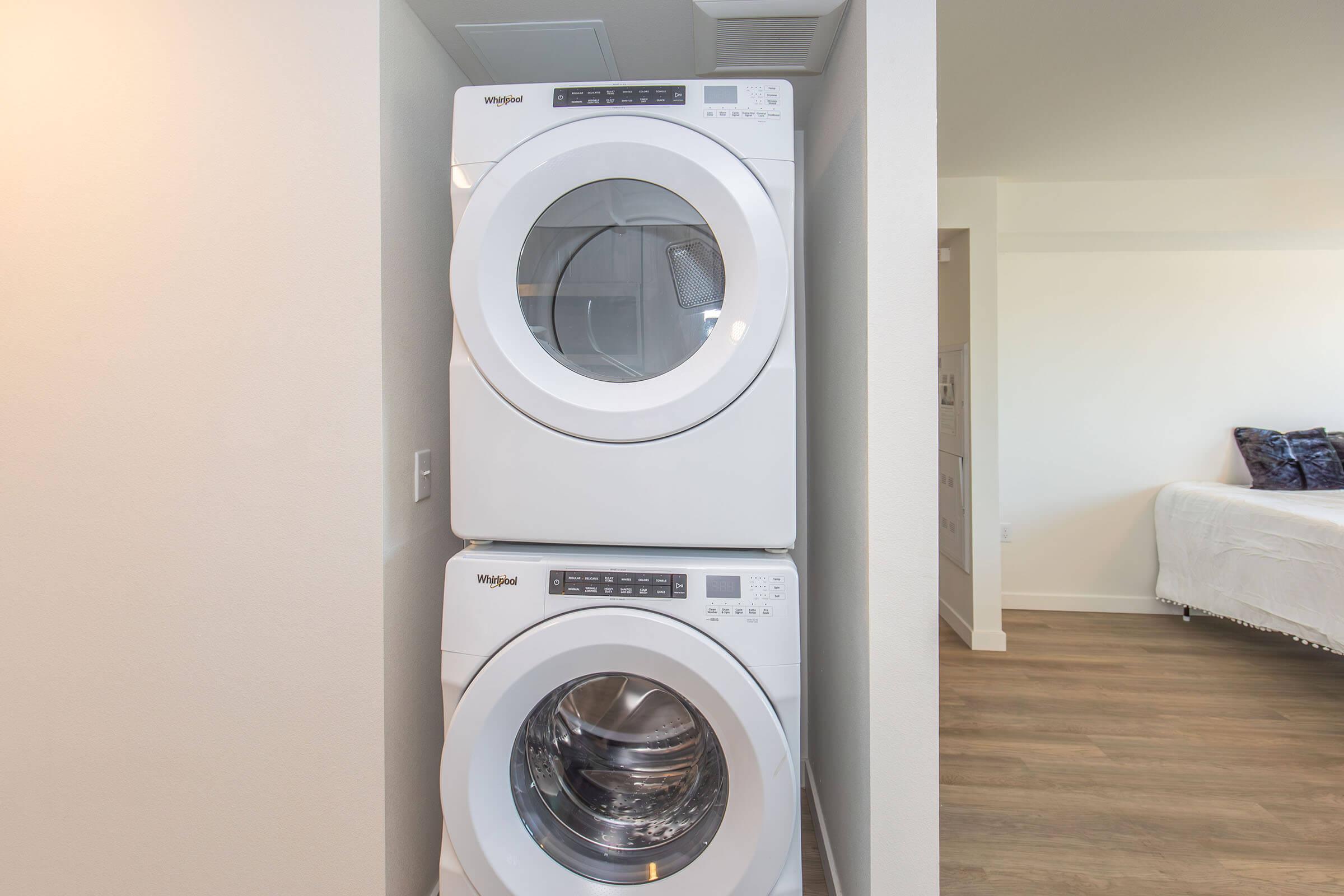 Stacked white washer and dryer units positioned in a corner of a modern interior space, featuring a clean design and visible control panels. The surrounding area has light-colored walls and wooden flooring, suggesting a contemporary laundry area.