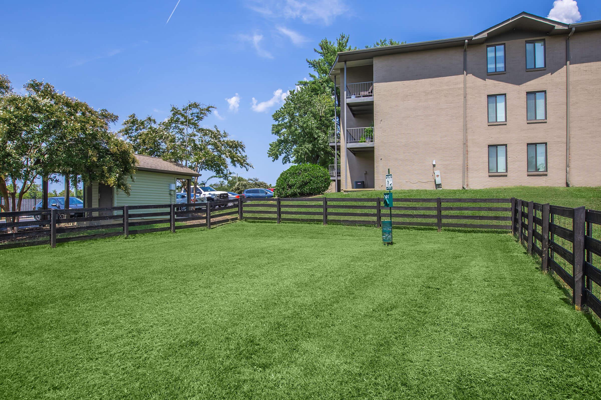 A green, well-maintained lawn area enclosed by black wooden fencing, situated next to an apartment building and a small shed. The scene is bright with blue skies and a few clouds, showcasing a tidy outdoor space suitable for relaxation or recreation.