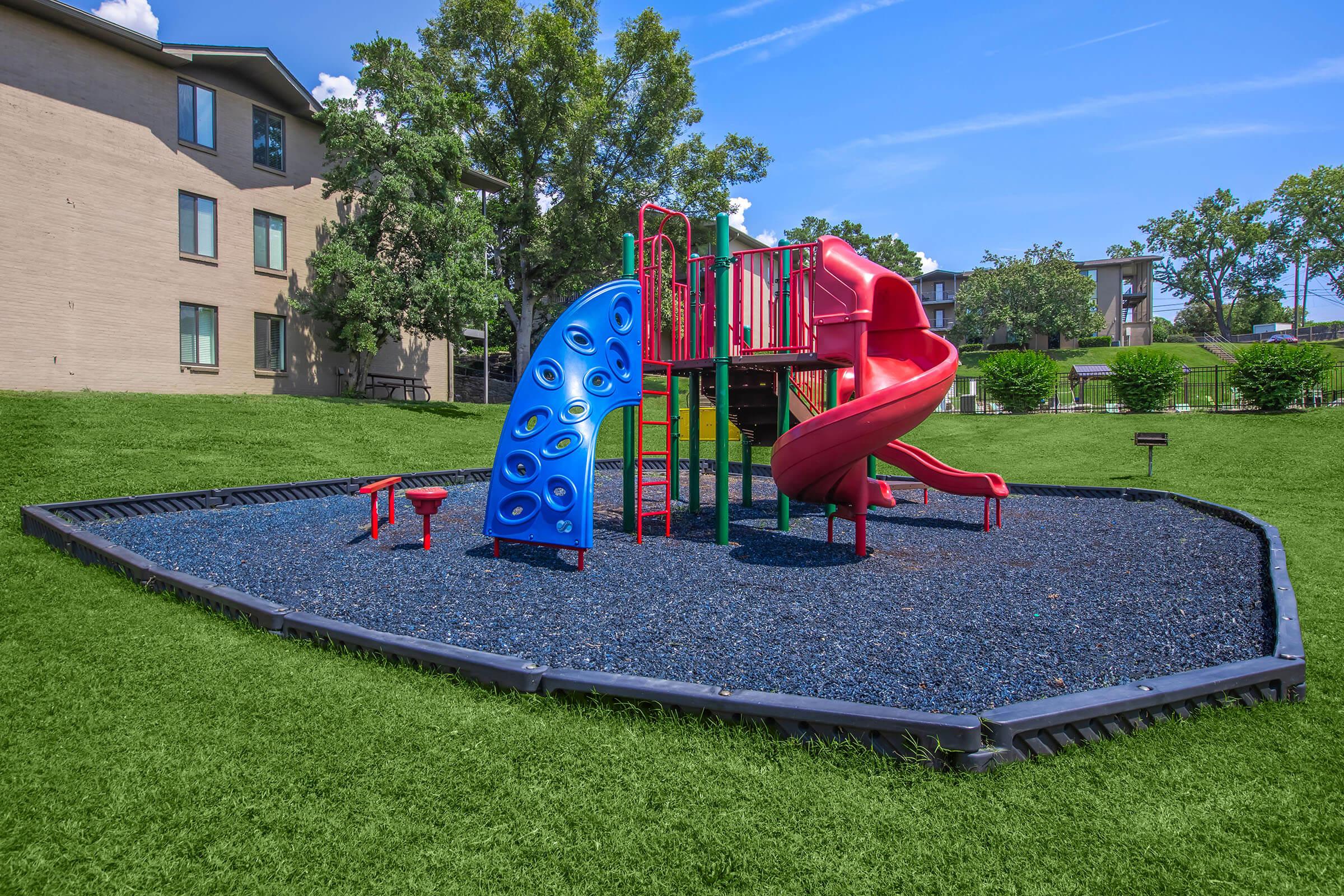 A colorful playground with a red slide, climbing wall, and play structures sits on green grass. The area features a soft, dark-blue ground cover for safety and is surrounded by trees and buildings. The sky is clear and bright, creating a cheerful outdoor play environment.