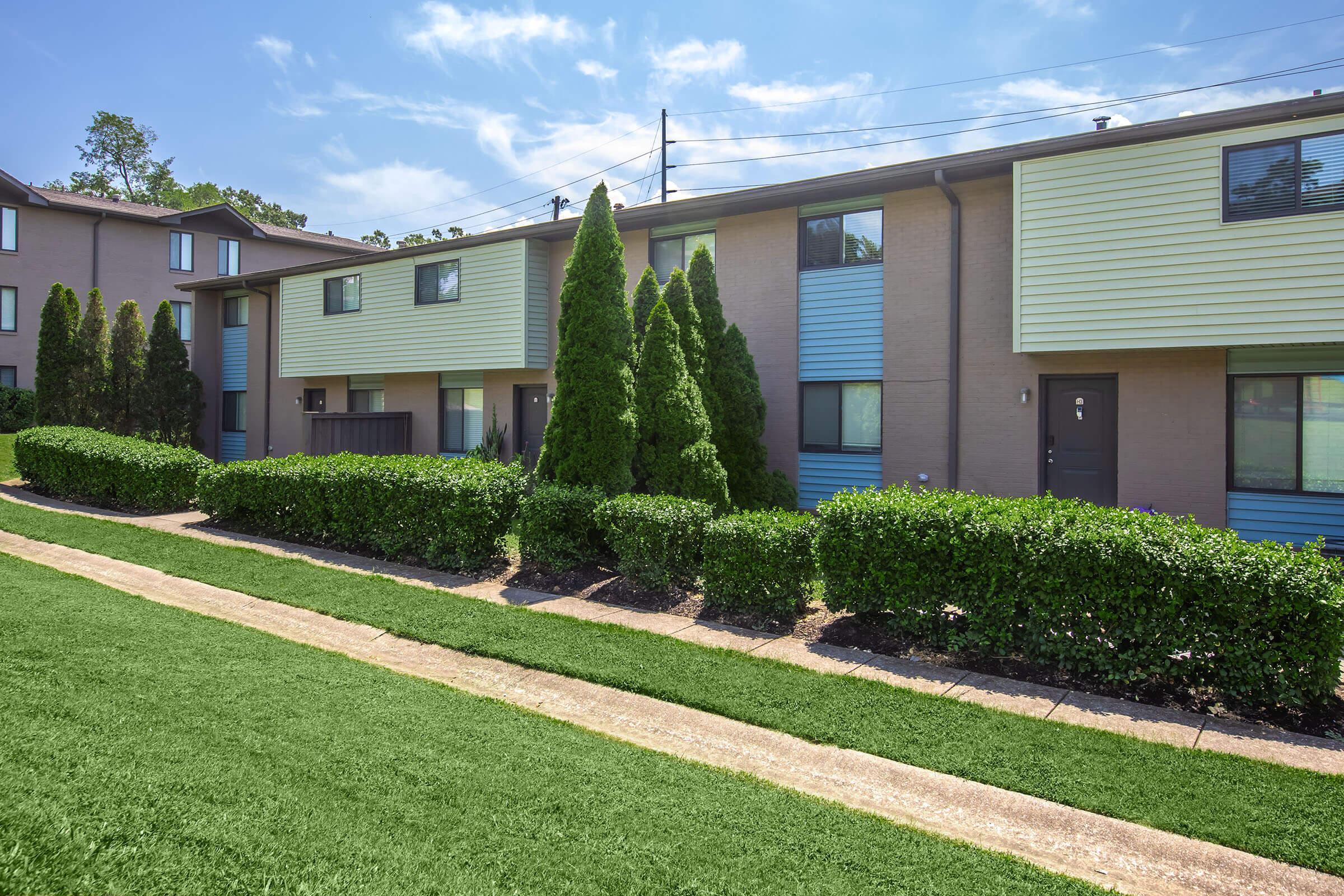 Row of modern townhouses with green shrubbery in front, a well-maintained pathway, and clear blue skies. The buildings feature a mix of beige and light blue siding with windows. Lush grass lines the pathway, creating a neat and inviting exterior.