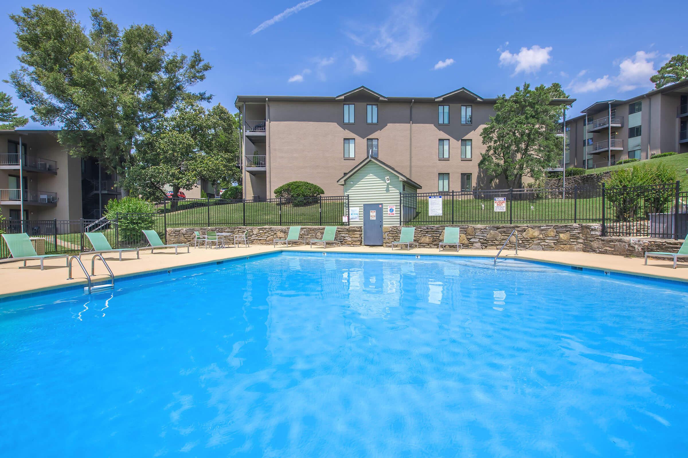 A bright blue swimming pool surrounded by green lounge chairs, with a stone border. In the background, there is a multi-story apartment building and well-maintained grass and trees. Clear blue sky with a few wispy clouds overhead.