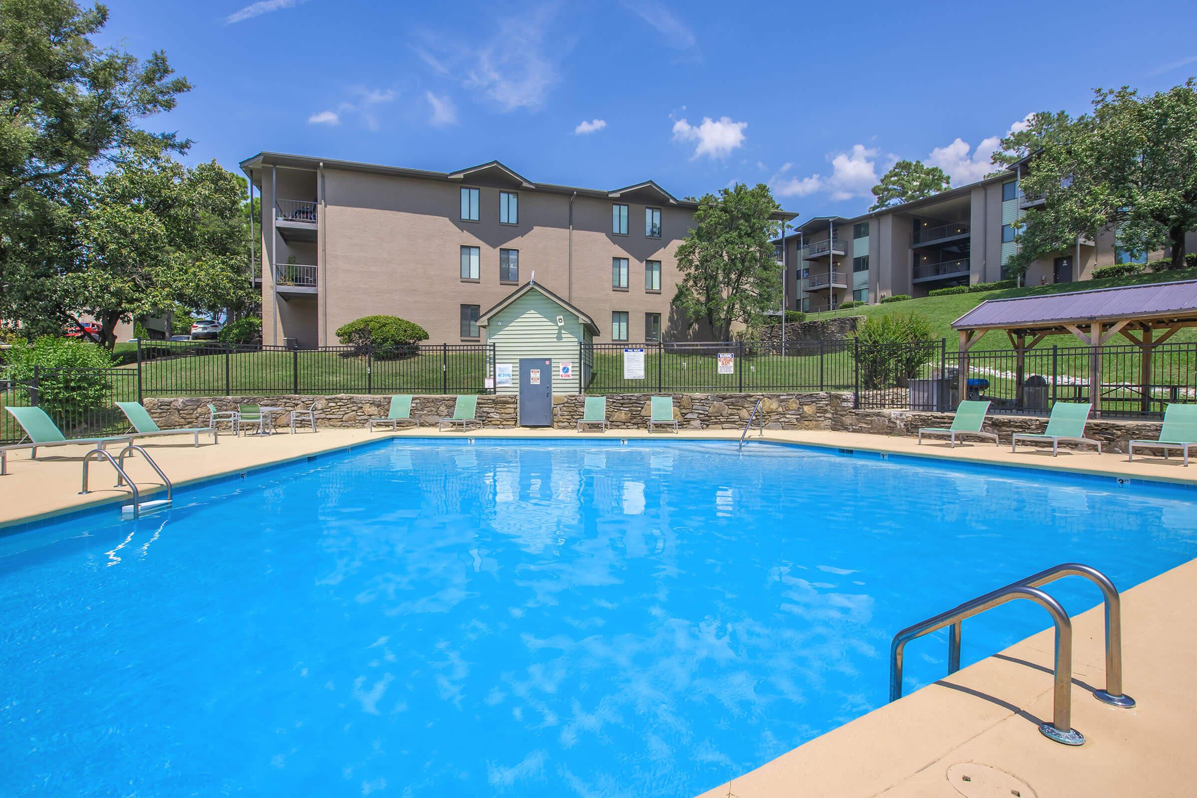 Swimming pool surrounded by lounge chairs and a stone wall, with a grassy area and apartment buildings in the background. The sky is clear blue with a few clouds, and there are trees lining the pool area.
