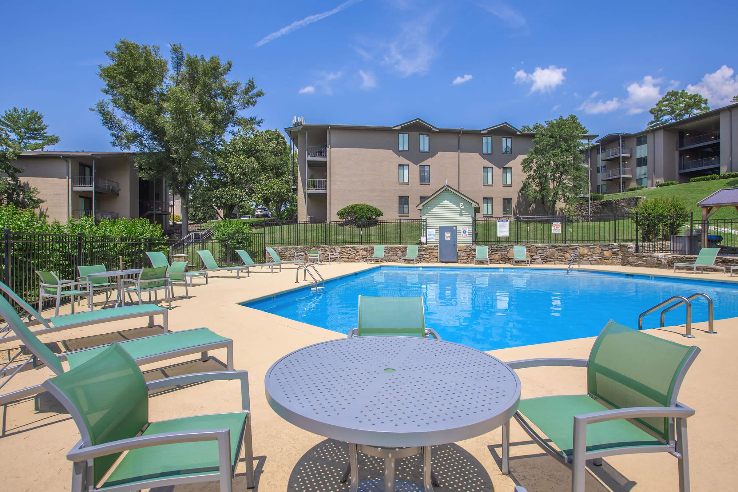 A clear blue swimming pool surrounded by green lounge chairs and tables. In the background, there are three apartment buildings and lush greenery. The sky is bright with a few clouds, creating a sunny and inviting atmosphere.