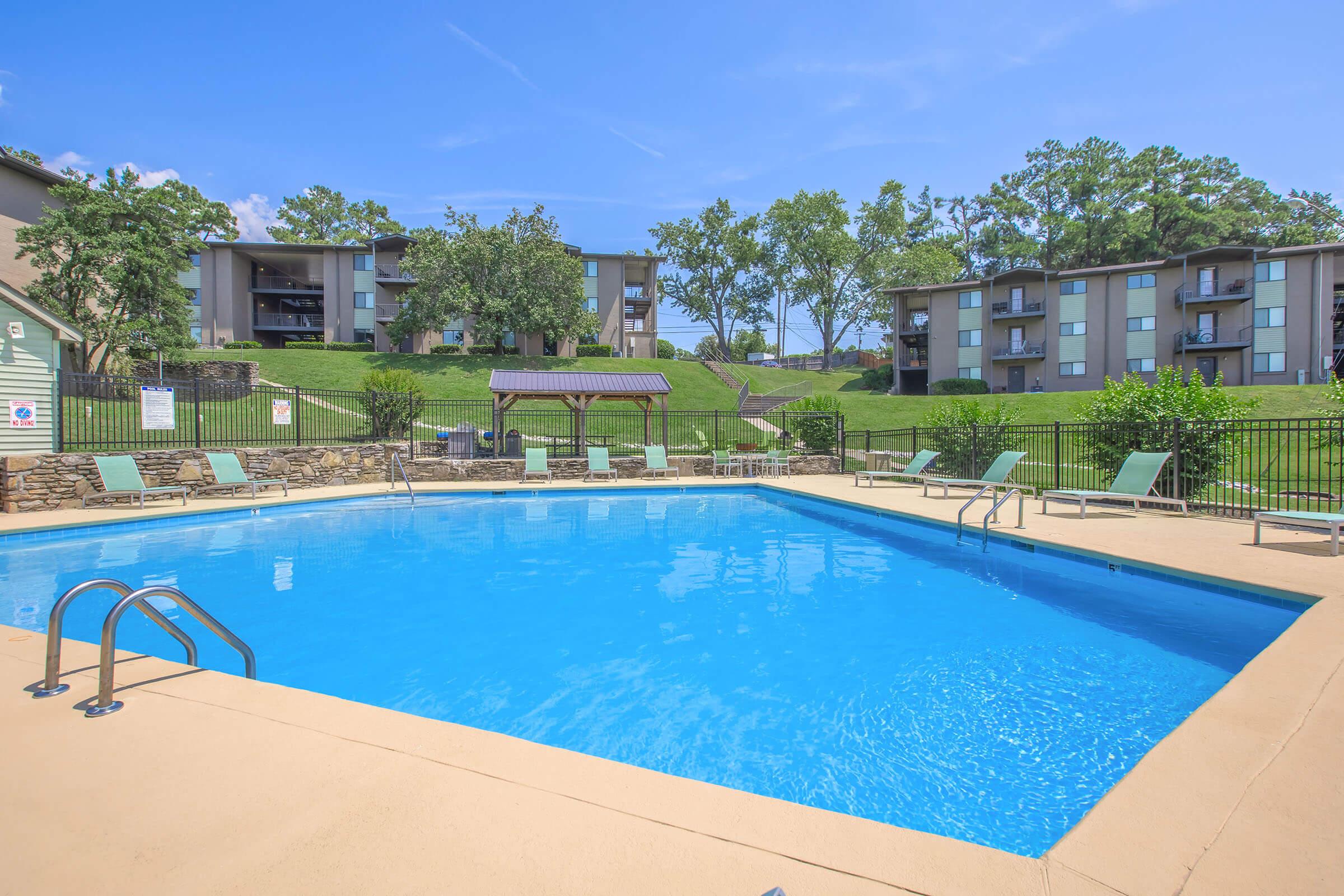 A clear blue swimming pool surrounded by lounge chairs, with green lawns and trees in the background. Several apartment buildings are visible on a sunny day. A shaded area with a picnic table is nearby, enhancing the outdoor leisure space.