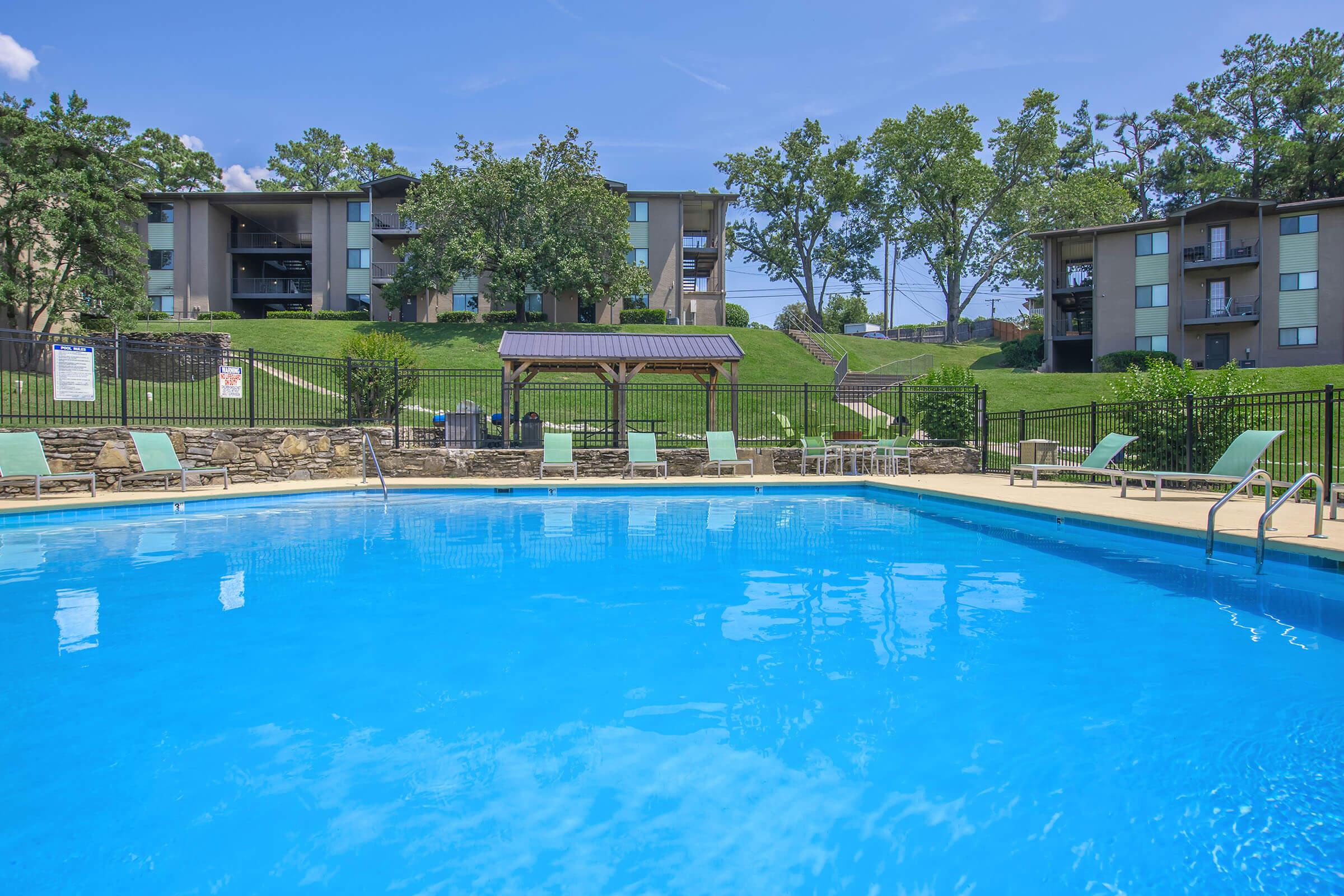 A sparkling blue swimming pool surrounded by lounge chairs, with a wooden gazebo nearby. In the background, there are green grassy areas and three multi-story apartment buildings under a clear blue sky with a few trees. The scene portrays a sunny, inviting outdoor space for relaxation and leisure.