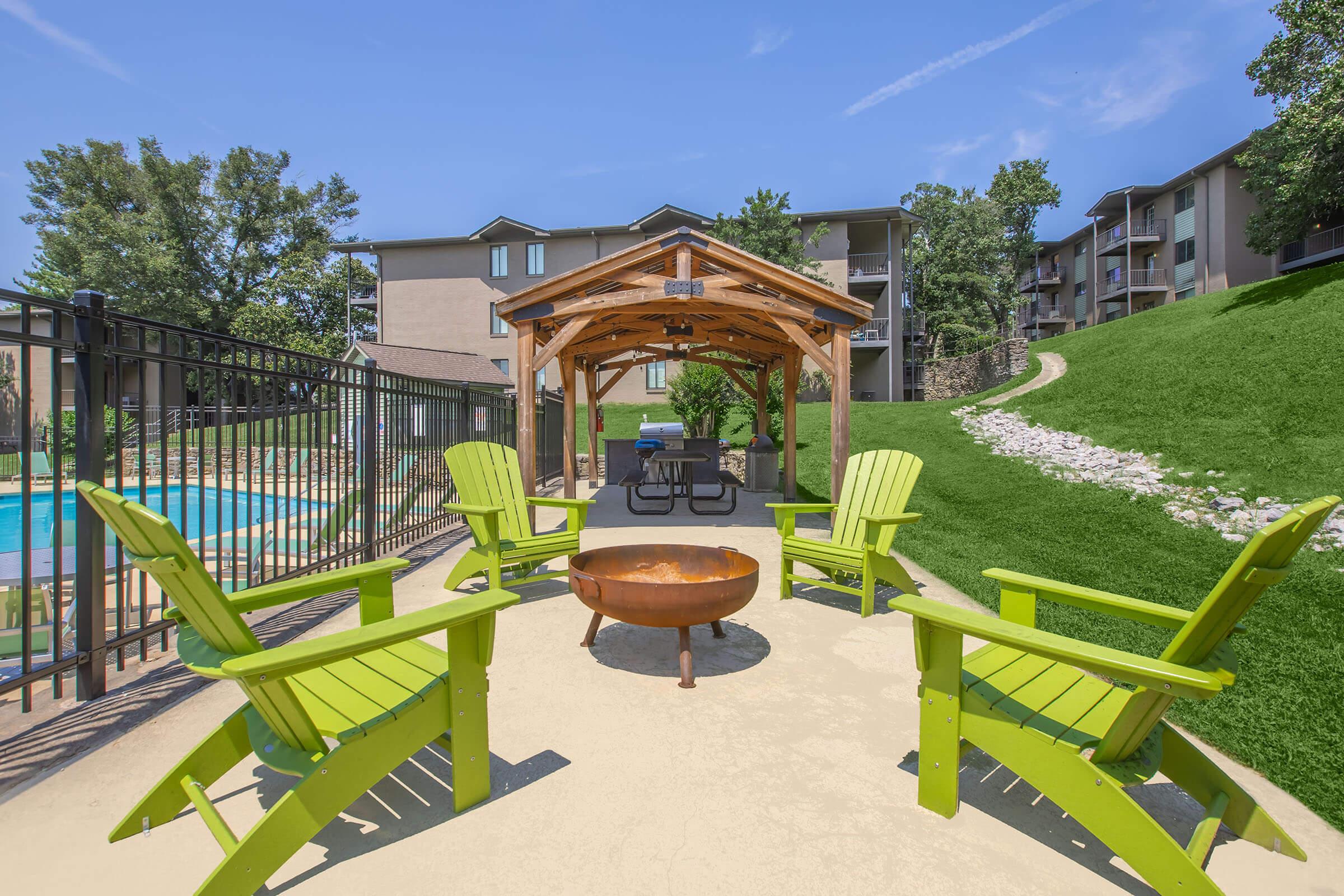 A serene outdoor area featuring bright green Adirondack chairs arranged around a circular fire pit. In the background, a swimming pool is visible, surrounded by a fenced area. A cozy wooden gazebo provides shade, with a picnic table inside. Lush greenery and apartment buildings frame the scene under a clear blue sky.
