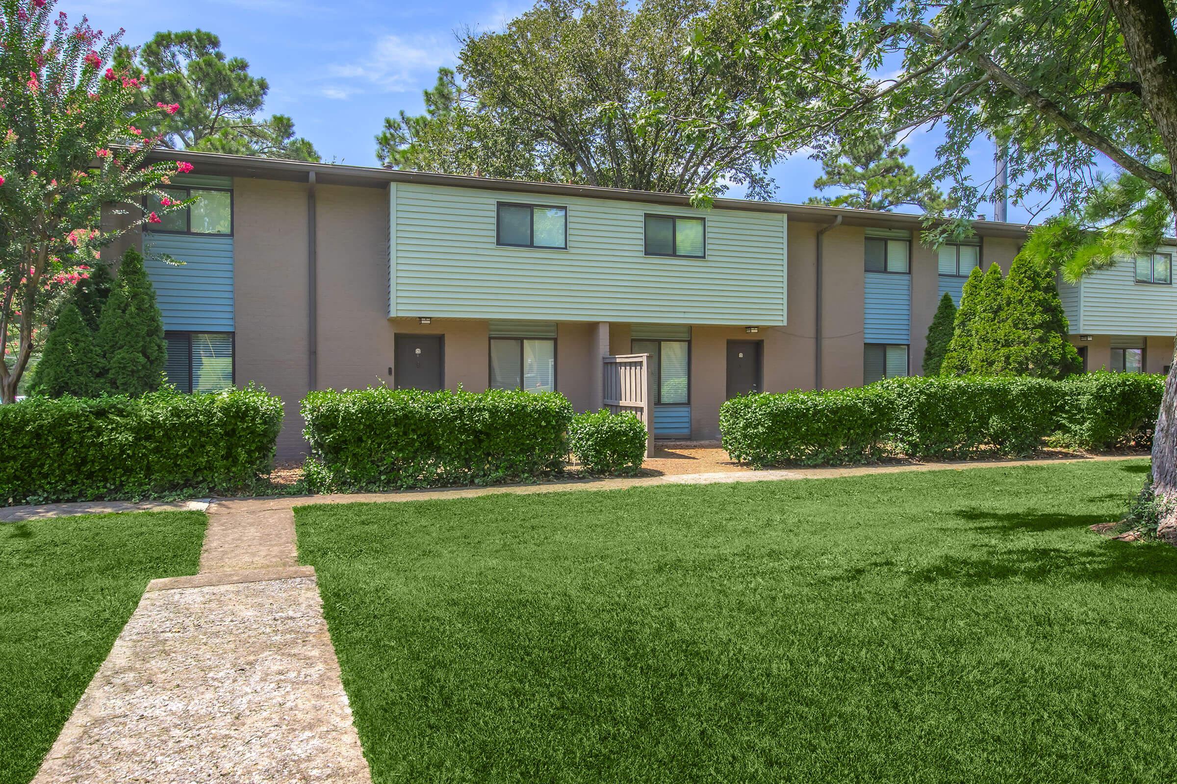 View of a landscaped apartment complex featuring two-story buildings with green siding. In front, a well-maintained lawn with manicured hedges and a stone walkway leading to the entrances of the apartments. The scene is surrounded by trees and a clear blue sky, creating a welcoming atmosphere.