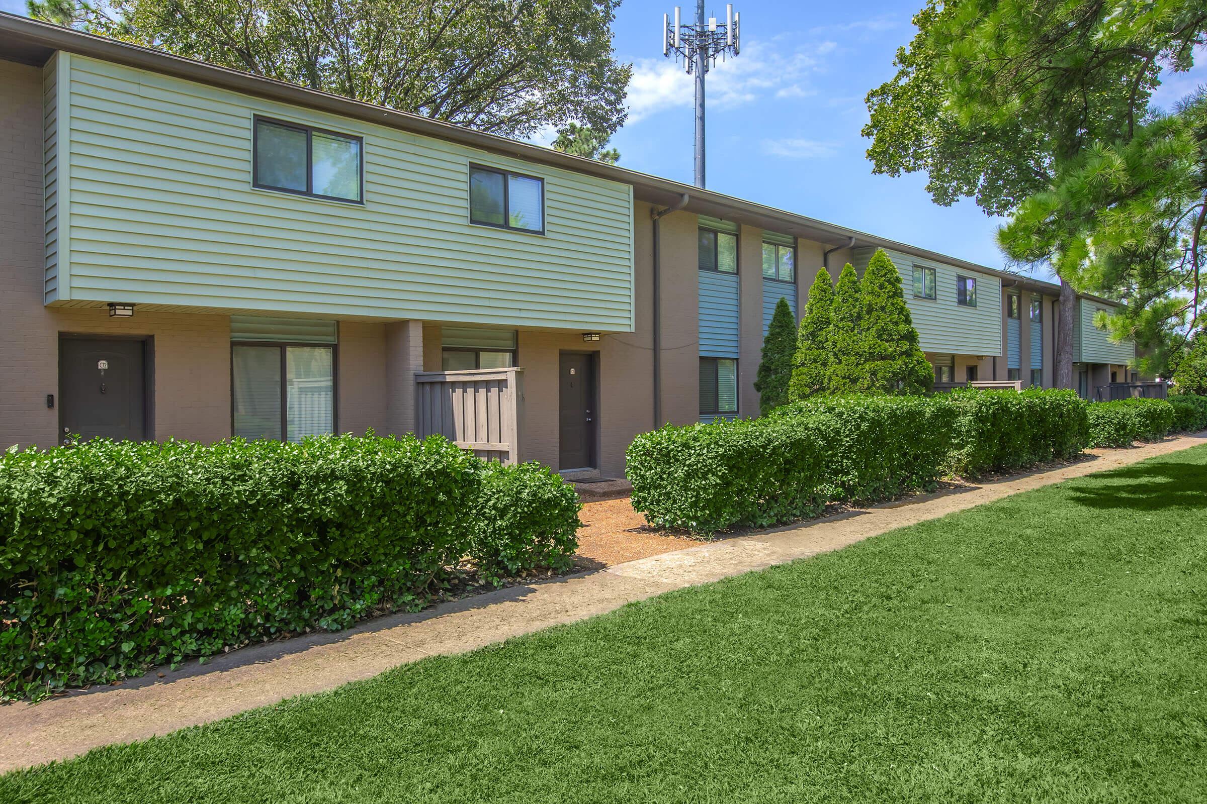 A row of two-story apartment buildings with a light green exterior. The buildings are lined with neatly trimmed hedges and small trees. A grassy area in the foreground and a cell tower visible in the background under a clear blue sky.